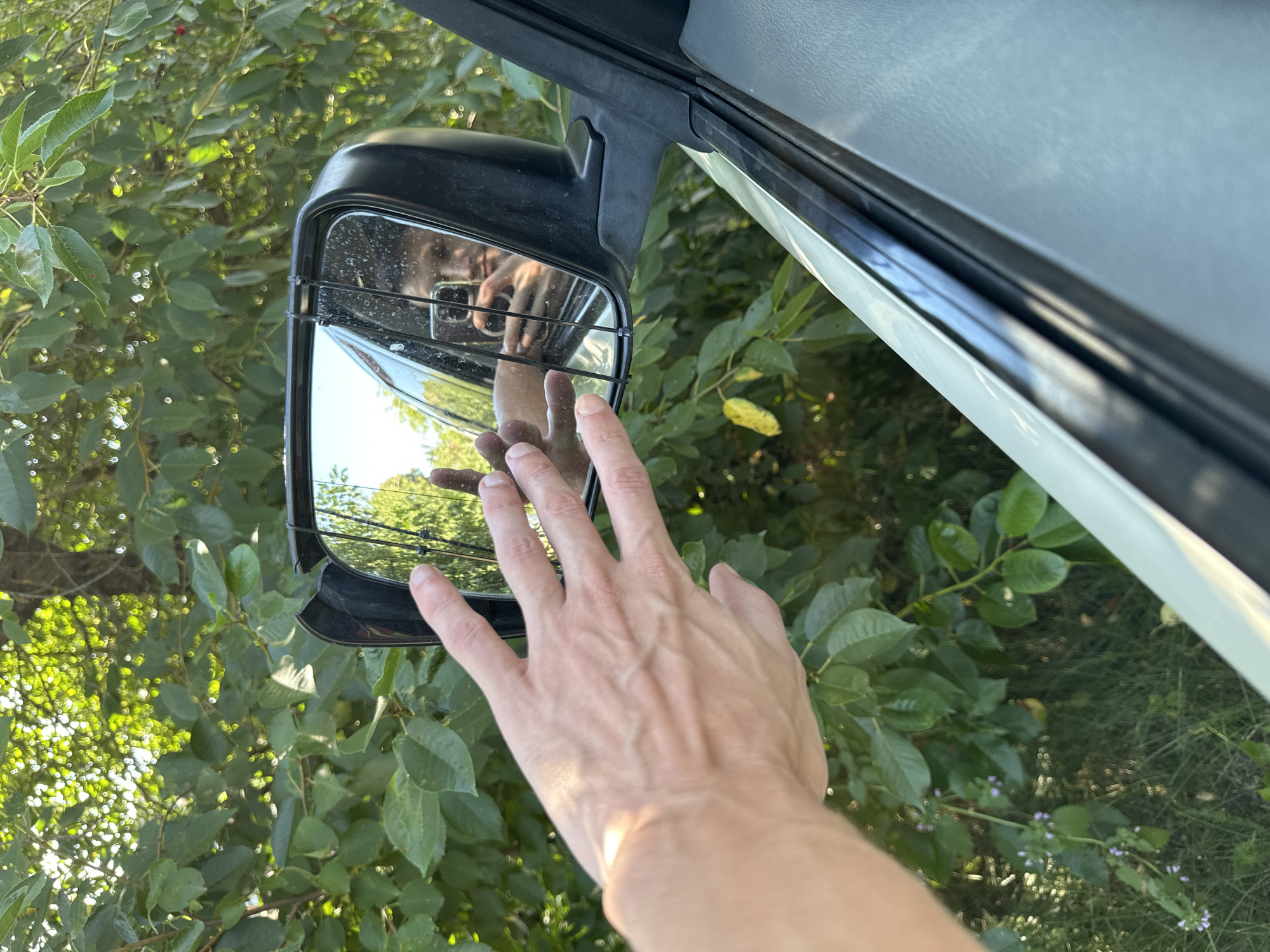 A person's left hand is extended towards a car's side mirror, its palm and fingers visible. The mirror reflects a person taking a selfie with a smartphone, holding it in their right hand, and looking at the phone's screen. The scene is set outdoors, with lush green foliage surrounding the vehicle. The car appears to be parked next to a tree-lined area, likely in a rural or suburban location. There is no indication that this scene is located in Shatava, Ukraine.