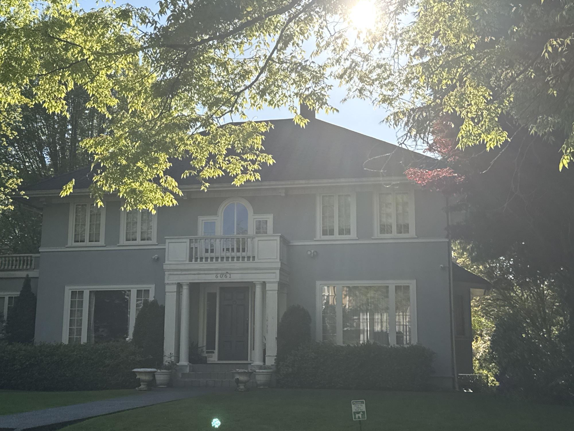 The image displays a stately two-story house with a light blue-grey exterior, set against a clear, sunny sky. The house is partially framed and obscured by abundant green foliage from large trees, through which bright sunlight streams, creating lens flare and illuminating the leaves from behind.
The house features a dark hip roof and numerous white-framed windows, some with multiple panes. A prominent front entrance is centered, showcasing a portico supported by two white columns, leading to a dark main door. Above the portico, on the second floor, is a white balcony with a balustrade railing, and an arched window is situated centrally above it. Another balcony with a similar railing extends from the left side of the second floor. The ground level includes large windows on either side of the entrance. Security cameras are visible on the house.
A well-maintained lawn extends in front of the house, with a dark pathway leading to the front steps. Decorative urns or planters are positioned on either side of the steps. Lush green shrubs and trimmed topiaries line the base of the house. Some reddish-brown foliage is visible near the right side of the house, possibly from a Japanese maple. The bright sunlight suggests it is either morning or late afternoon.
The house number "6061" is clearly visible above the front door. On the lawn, a small white sign with black text reads "Please Clean Up After Your Dog," accompanied by a dog graphic. There is no human activity present in the scene.