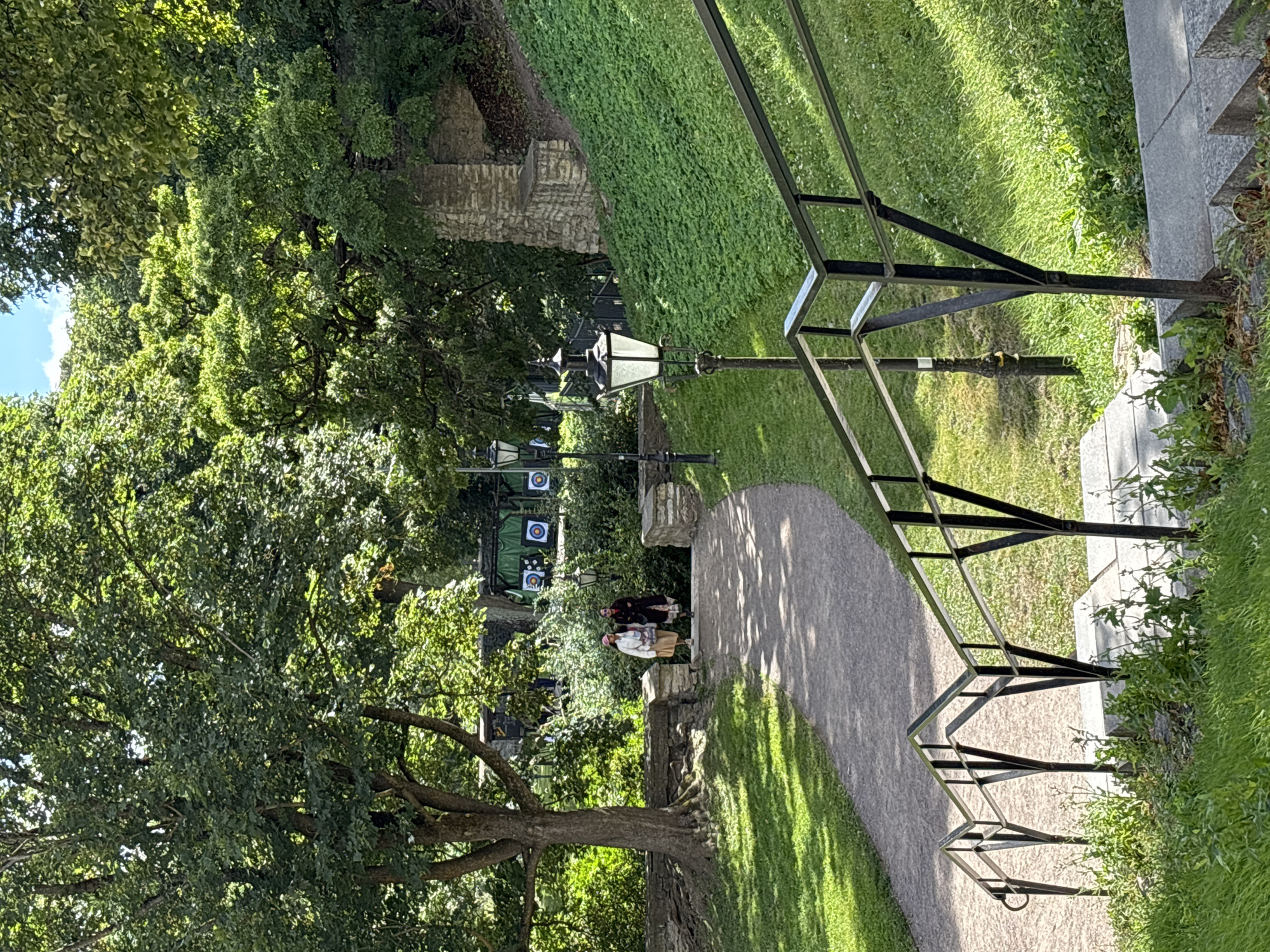 The image depicts a daytime scene in a park setting within Tallinn, Estonia. In the foreground, a stone staircase with a black metal railing leads upwards. To the left of the stairs, a path winds through a grassy area shaded by large trees. Further up the path, two individuals, appearing to be women, are walking side by side. One is wearing a light-colored outfit with a hat, and the other is in darker attire. In the background, several archery targets are visible, suggesting an outdoor recreational area. The environment is lush with green trees and grass, indicating a pleasant, possibly summer, day. The overall atmosphere is serene and offers a glimpse into a public space in the city.