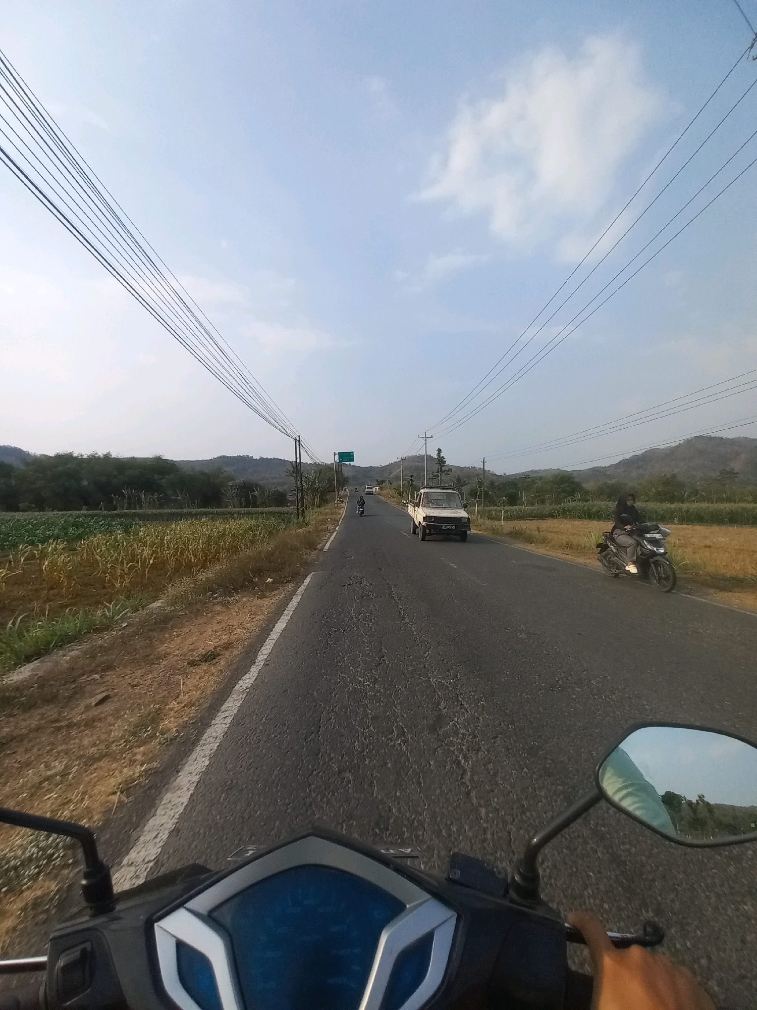 The image captures a view from the perspective of someone riding a scooter on a paved road in Kulurejo, Indonesia. The road stretches into the distance, bordered by dry, brown vegetation and a field of corn on the left. On the right side of the road, a person is riding a scooter, also heading away from the camera. Further down the road, a white truck is visible, and in the background, rolling hills covered with trees rise against a clear sky with scattered clouds. Overhead, multiple power lines run parallel to the road. The foreground shows the handlebars and dashboard of the scooter the photo is being taken from.