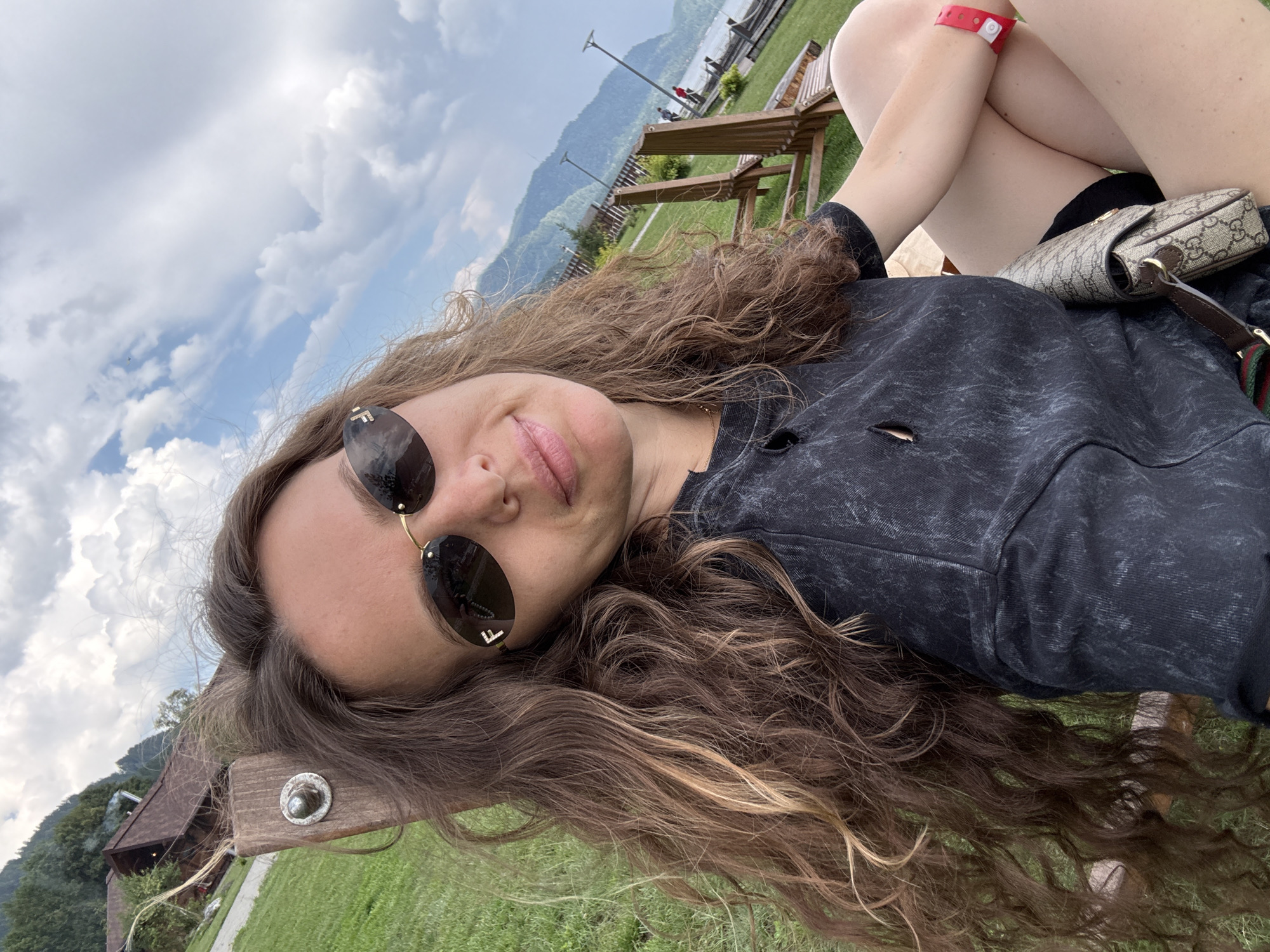 A woman with long, wavy brown hair and sunglasses is sitting on a wooden lounge chair outdoors. She is wearing a dark, t-shirt-style top and a small crossbody bag with a monogram print. A red wristband is visible on her left wrist. The background shows a grassy slope with trees and a body of water, likely Lake Teletskoye, with mountains in the distance. A wooden building with a brown roof is also visible on the left side of the frame. The sky is partly cloudy. The location is identified as the village of Artybash, Russia.