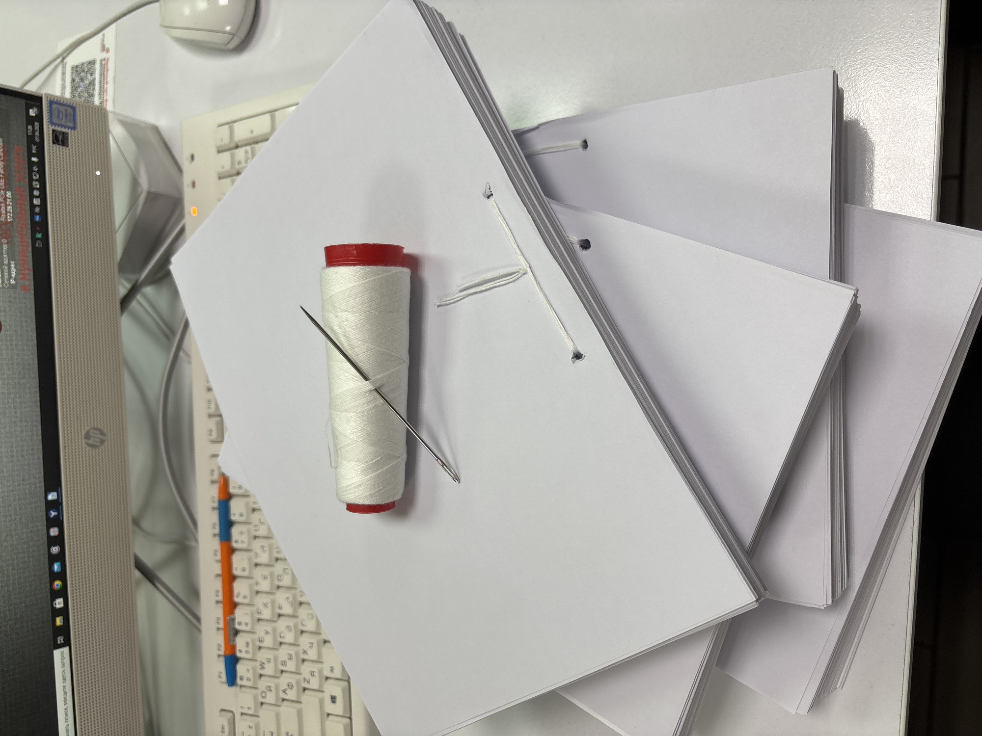 A top-down, brightly-lit shot depicts a stack of blank, white, hole-punched papers stacked somewhat haphazardly on a white desk surface, close to a white keyboard and a light gray computer monitor. A bobbin of white thread is placed on top of the topmost sheet of paper with a needle stuck into it. The desk and objects are likely located in an office or study in the city of Chaikovsky, Russia.
