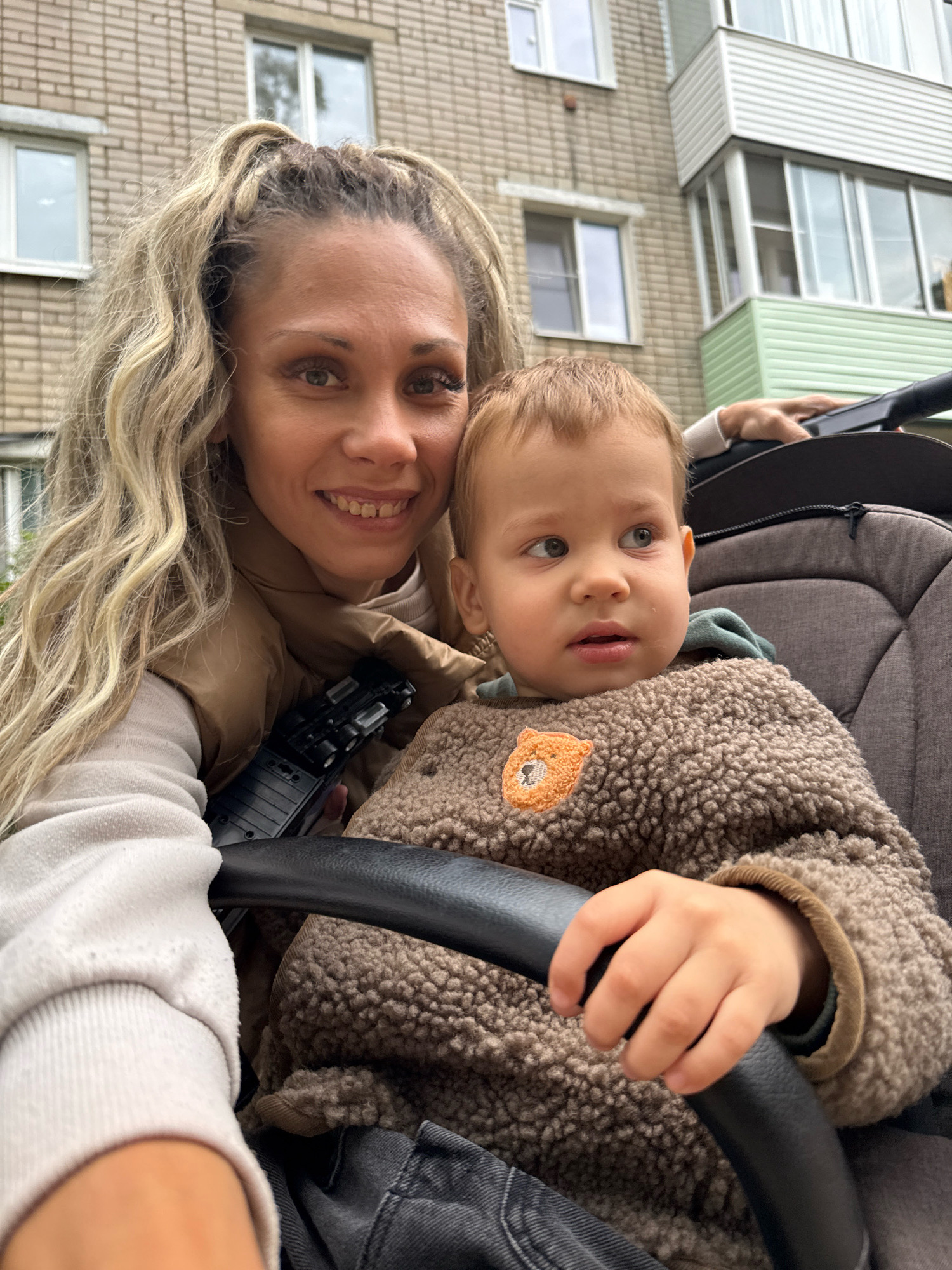 A woman with blonde, wavy hair and a tan vest smiles at the camera, holding a baby in a stroller. The baby, wearing a brown, fuzzy jacket with a bear patch, looks to the side. They are outdoors, with a brick building and a balcony visible in the background, suggesting a location in the city of Rusanovo, Russia. The overall scene appears to be a candid moment captured in an urban environment.