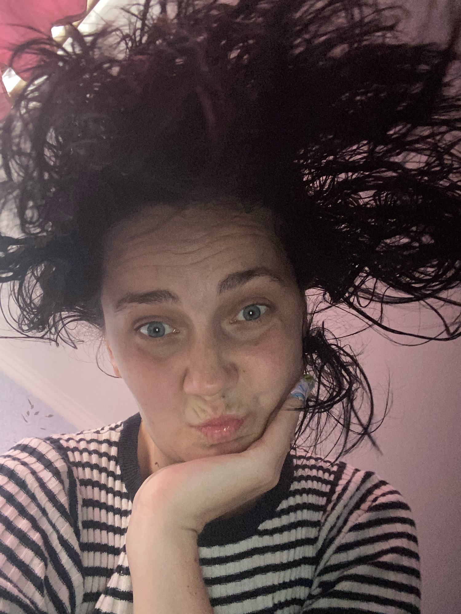 A close-up, low-angle, indoor, selfie-style photograph shows a young woman with very messy, dark, curly hair that stands out from her head. She is wearing a black and white striped long-sleeved shirt. Her left hand is propped under her chin, and she is looking directly at the camera with a slightly pouting expression. Her eyes are blue and wide open. The background is a pink wall with some faint patterns. The lighting is dim and appears to be artificial, casting a slight pink hue on the scene. The overall impression is one of a casual, spontaneous moment captured in an unknown indoor location.