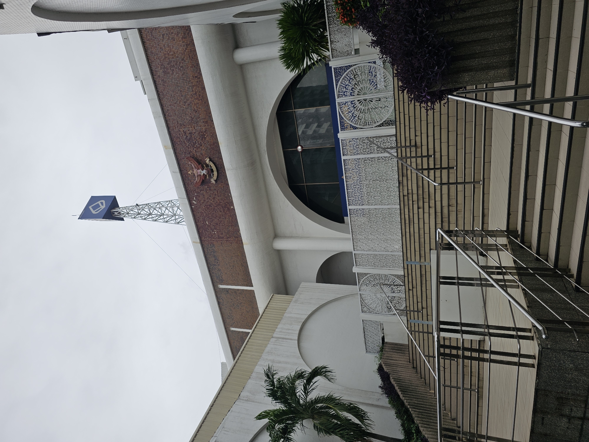 This is a medium-angle, exterior shot of a building entrance in Bangkok, Thailand. The building features a white facade with large arched windows and ornate white decorative panels. A set of wide, outdoor stairs with metal railings leads up to the entrance. To the left of the stairs, a palm tree is visible. Above the building, a tower with a blue sign displaying a white logo is topped with a metallic structure. A national emblem is mounted on the brick portion of the building. The sky is overcast, suggesting a cloudy day.
