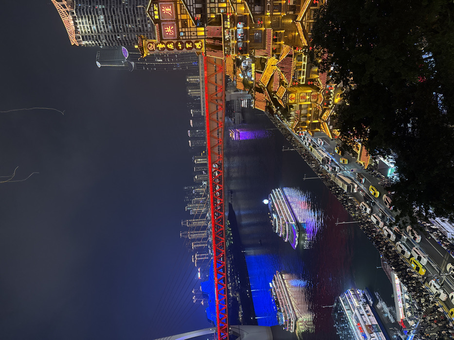 This is a nighttime aerial view of the Hongyadong commercial complex and surrounding area in Yuzhong District, Chongqing, China. The scene depicts a bustling urban waterfront environment.
In the foreground and mid-ground on the right, the multi-tiered Hongyadong complex is brightly illuminated with golden, red, and orange lights, showcasing traditional stilt-house architecture extending vertically from the riverbank. To its left, a heavily trafficked road runs parallel to the river, featuring numerous vehicles including cars, buses, and yellow taxis, alongside dense crowds of pedestrians lining the promenade.
The dark body of water, identified as either the Jialing or Yangtze River, occupies the center-left. Several large cruise boats or ferries, elaborately illuminated with multi-colored lights (blue, purple, white), are visible navigating the river, reflecting their illumination on the water's surface.
Spanning the river in the upper mid-ground is the Dongshuimen Bridge, a cable-stayed bridge. Its main truss structure is distinctly illuminated in red, while its cables and support tower on the left are lit with blue light.
In the background, beyond the bridge and across the river, a dense skyline of numerous modern high-rise buildings with illuminated windows forms the distant urban landscape under a dark night sky. Thin, indistinct lines are visible in the upper sky, likely light reflections or atmospheric phenomena. The overall scene is characterized by extensive artificial lighting and a high level of human and vehicular activity.