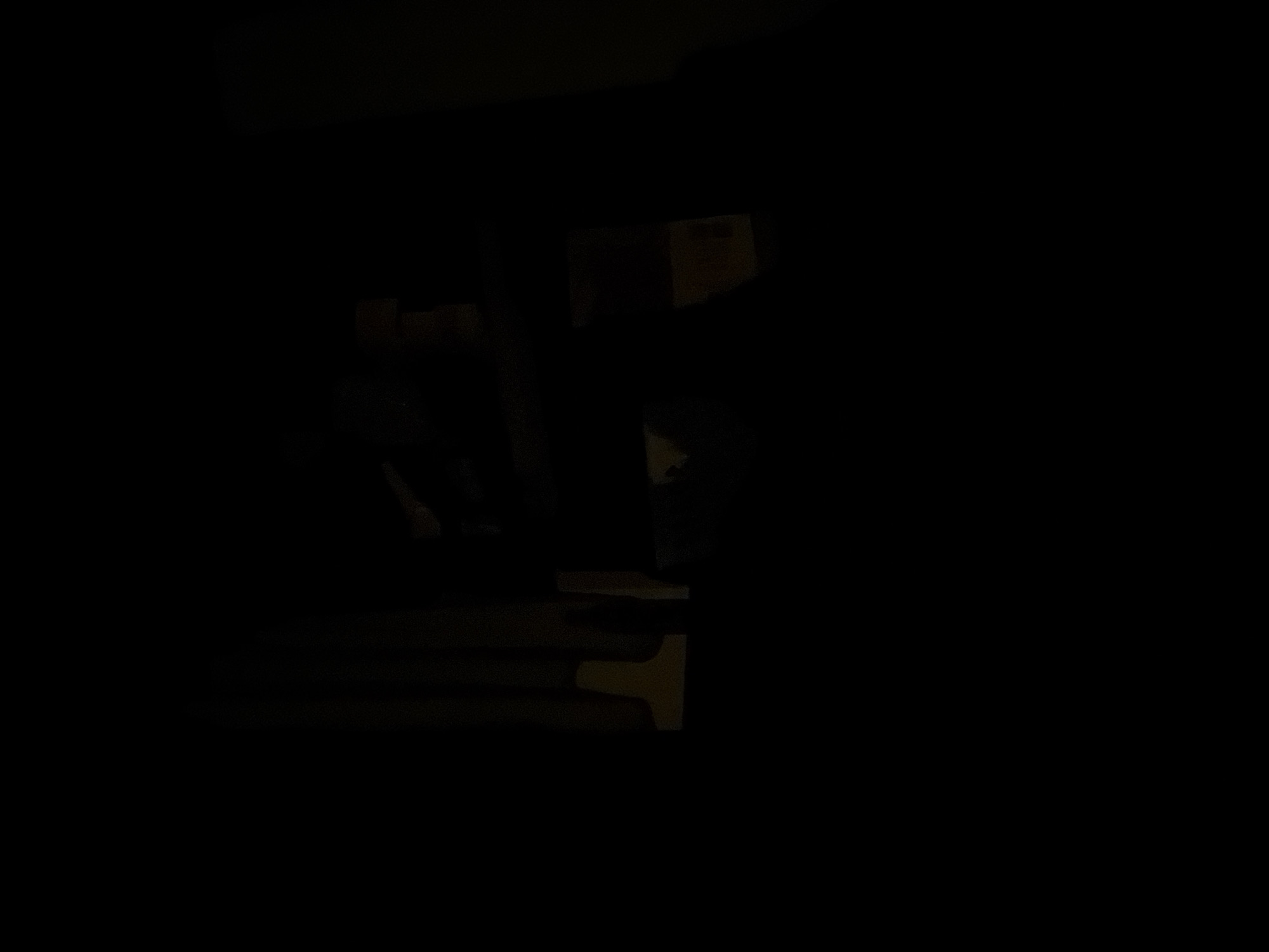 The image is a dimly lit indoor scene with very low visibility. The majority of the image is black, suggesting the absence of significant light sources or the presence of heavy shadows. In the upper central portion of the image, there are faint outlines of objects that appear to be stacked or arranged on a surface. These objects are indistinct and their nature cannot be definitively identified due to the poor lighting. Some of these shapes suggest rectangular forms, possibly boxes or containers, with varying textures or colors that are barely discernible. There are also vertical lines visible, which could indicate curtains or the edge of furniture. The overall impression is of a dark, possibly cluttered, indoor space where details are obscured.