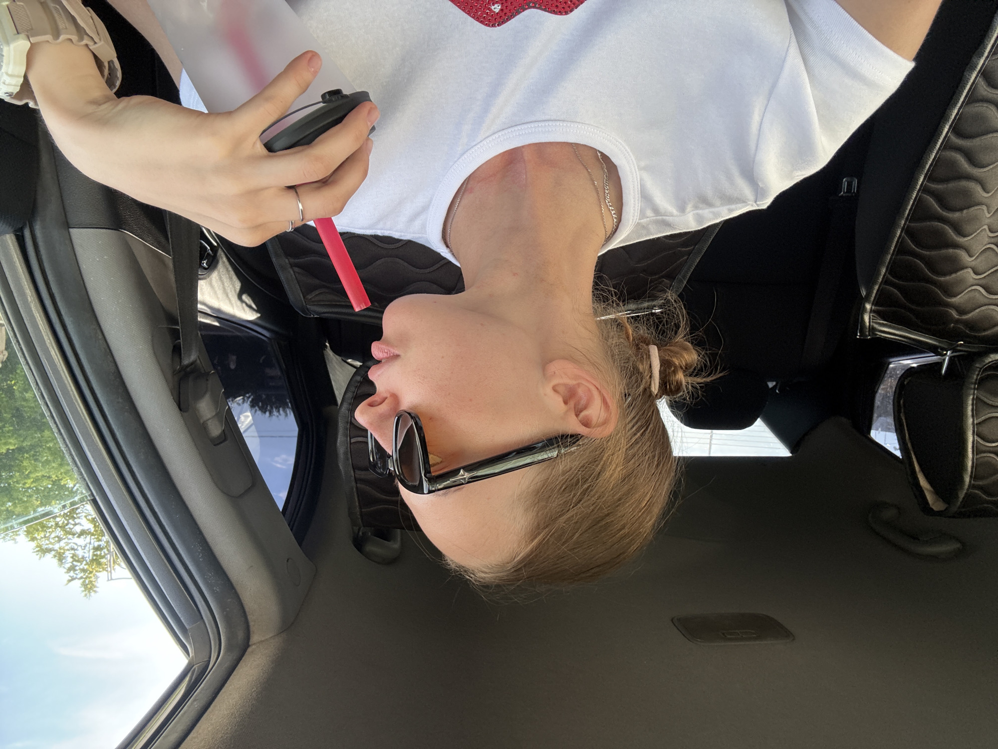 In an indoor, side-facing shot, a woman with blonde hair pulled into a bun, wearing sunglasses and a white t-shirt, sits in the passenger seat of a car. She is holding a clear plastic cup with a red straw in her right hand, bringing it to her lips. The interior of the car is visible, including the black seats and headrests, and a portion of the dashboard. The exterior of the car reveals trees and a bright blue sky through the open window. The setting is not specific to the city of Perm, Russia.