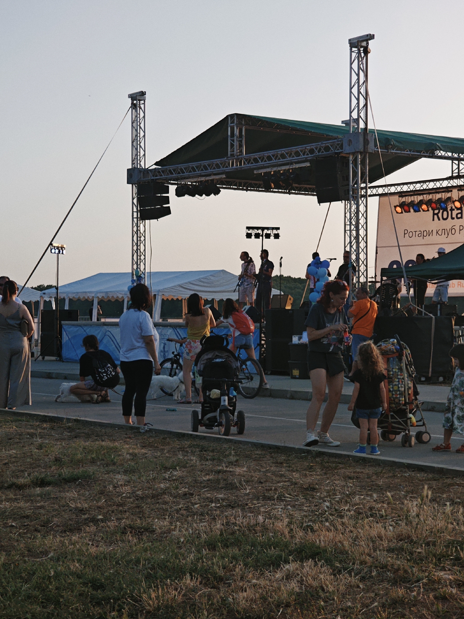 This media file captures an outdoor event with a live band performing on a stage. The scene is populated with a diverse group of people, including adults and children, interacting and enjoying the music. Several individuals are standing and observing the performance, while others are seated on the ground. Children are visible, with some accompanied by adults and others near strollers and a bicycle. The presence of dogs suggests a relaxed, family-friendly atmosphere.
The event appears to be taking place in the late afternoon or early evening, as indicated by the warm, soft lighting and long shadows. The sky is clear and bright, suggesting fair weather. The setting includes a large stage structure with scaffolding, lighting equipment, and speakers. Behind the stage, tents are set up, possibly for vendors or additional event facilities. A banner is displayed on the stage, featuring the text "Rotary Club P" in large letters and "Ротари клуб Р" below it.
The overall mood seems to be one of enjoyment and community gathering, with people of various ages engaging with the event and each other. The activity taking place is clearly a live musical performance, with band members visible on stage. The scene is set on what appears to be a paved area, with grassy areas surrounding it, hinting at a park or public outdoor space.