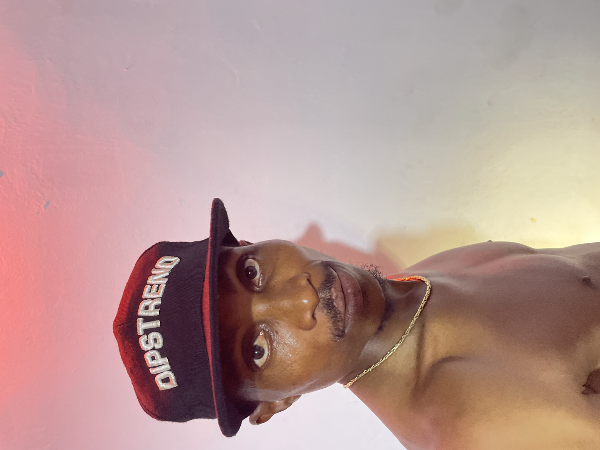 A man wearing a black baseball cap with the embroidered text "DIPSTREND" in white is looking directly at the camera. He has a thin mustache and a goatee. He is shirtless, revealing a gold chain around his neck. The background is a plain, lightly textured white wall with a soft red glow at the top, possibly from overhead lighting. The overall lighting is warm and somewhat diffused. The image appears to be a self-portrait or casual snapshot. The specific location in Adankolo, Nigeria, is not discernible from the visual information alone.