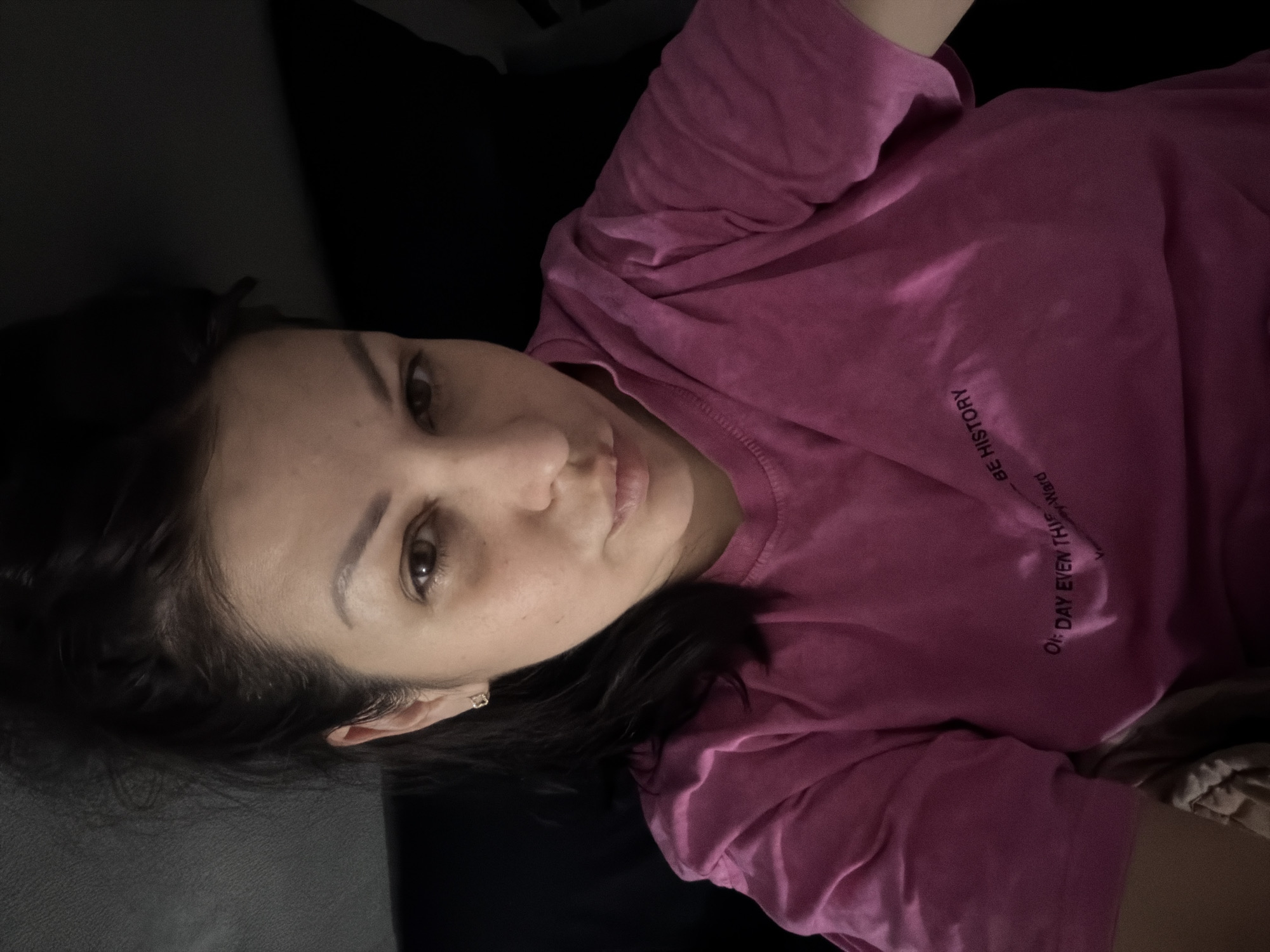 A woman with dark hair is pictured from the chest up, looking directly at the camera. She is wearing a pink t-shirt with text printed on it. The text on her shirt reads, "OH, DAY EVEN THIS" and "BE HISTORY." The woman has dark hair pulled back and is wearing a small, gold stud earring. The background is dark and appears to be indoors, possibly in a car or on a couch. The image was taken in Sochi, Russia.