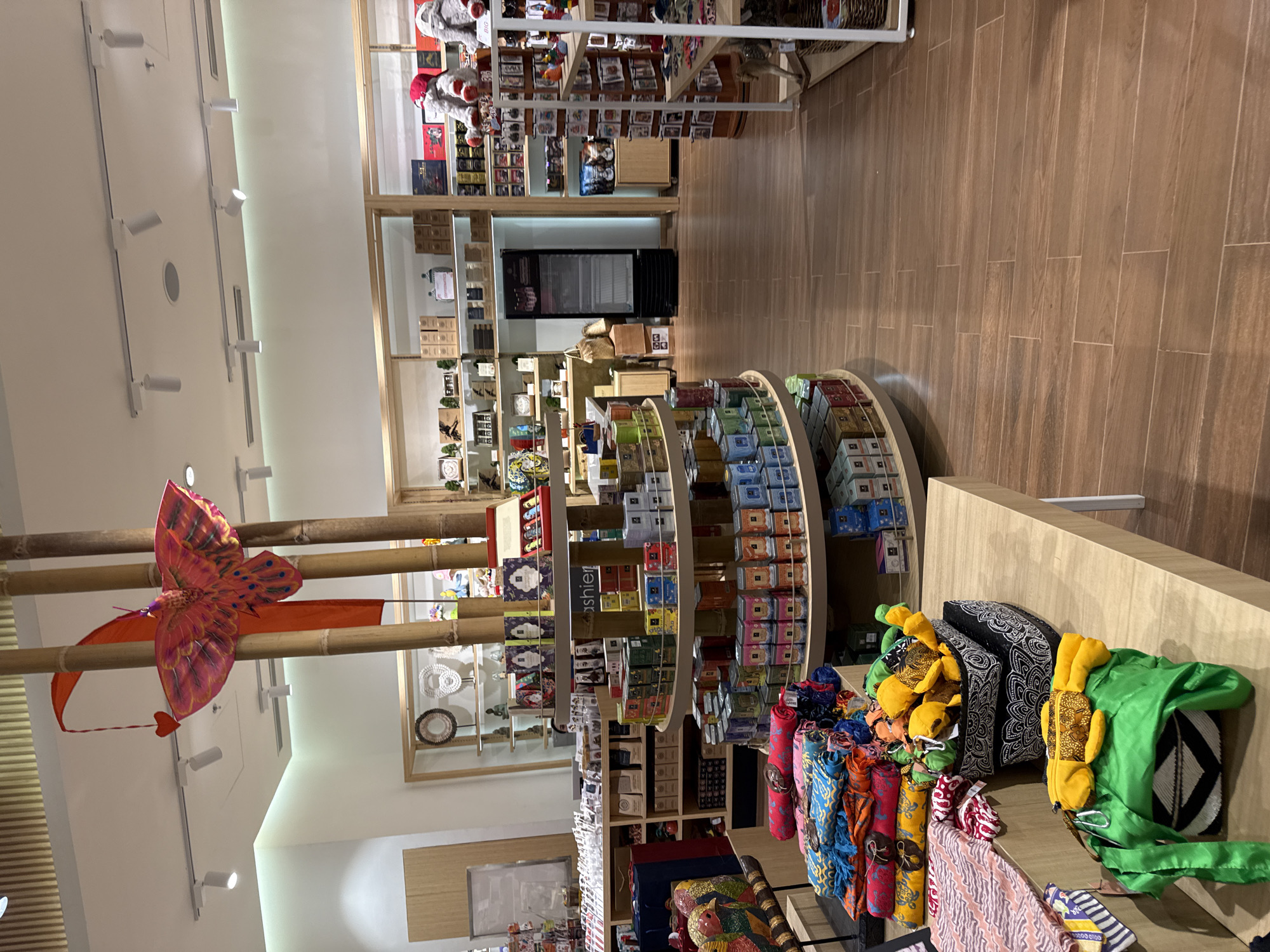 This is a wide-angle, eye-level shot of the interior of a gift shop in an unknown city in Indonesia. The shop features a variety of merchandise displayed on shelves and circular racks. Prominently featured in the foreground is a wooden counter displaying colorful textiles and woven baskets filled with yellow plush toys. Behind this, a large circular display rack is filled with various packaged goods, likely food items or small gifts, in colorful tins and boxes. Further back, shelves line the walls, filled with more packaged goods, decorative items, and some larger items like teddy bears. A prominent display of hanging bamboo poles with an orange kite and a red heart decoration hangs from the ceiling, adding a decorative element to the space. The floor is covered in polished wooden planks. In the background, a large black refrigerator is visible, and a checkout counter with a "Cashier" sign is also present. The lighting is bright and even, consistent with an indoor retail environment. There are no people visible in the image.