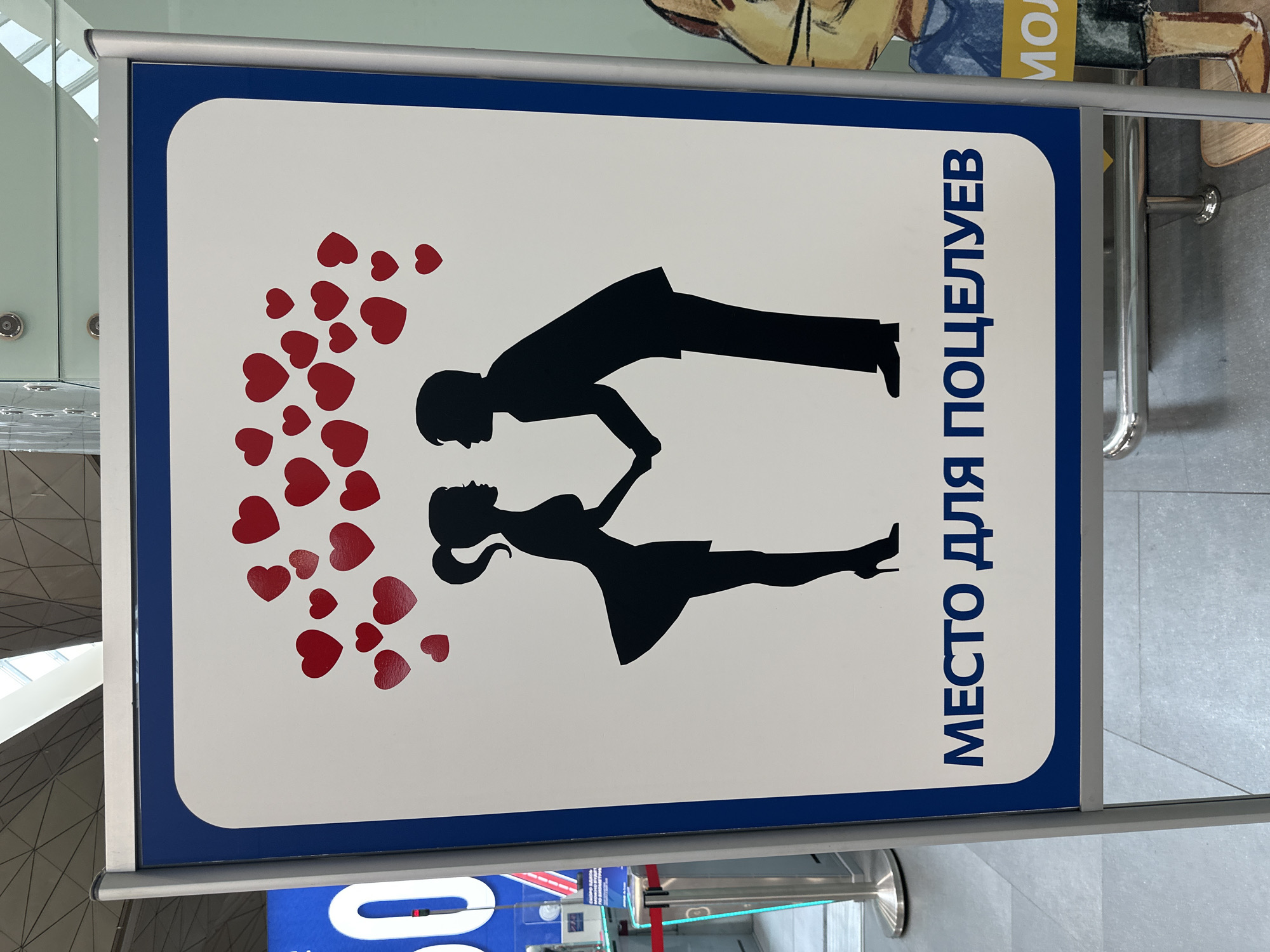 The image depicts a sign, likely at an airport, that features a white background with a blue border. A black silhouette of a man and woman in an embrace is positioned centrally on the sign, accompanied by numerous red heart shapes radiating from them on the left side. The Russian text "МЕСТО ДЛЯ ПОЦЕЛУЕВ" (MESTO DLYA POTSELUYEV) is printed vertically in blue on the right side, which translates to "Kissing Spot" or "Place for Kisses".
The location is likely Saint Petersburg, Russia, considering the Russian language.