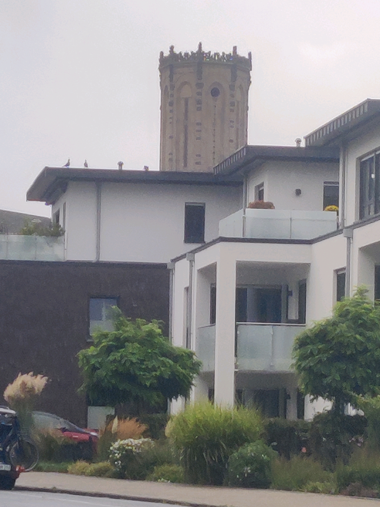 The image shows a residential building with multiple stories and balconies, in front of which are some trees and bushes. A distinctive tower structure is visible in the background, which is a prominent landmark in Emmerich on the Rhine, Germany. The sky is overcast, suggesting a cloudy day. There are no people visible in the image. The building appears to be modern, with white facades and glass railings on the balconies. The tower in the background is made of stone and has a unique architectural style, with arched windows and decorative elements. The foreground shows a street or sidewalk, with a few plants and what appears to be a motorcycle or scooter parked on the left side.