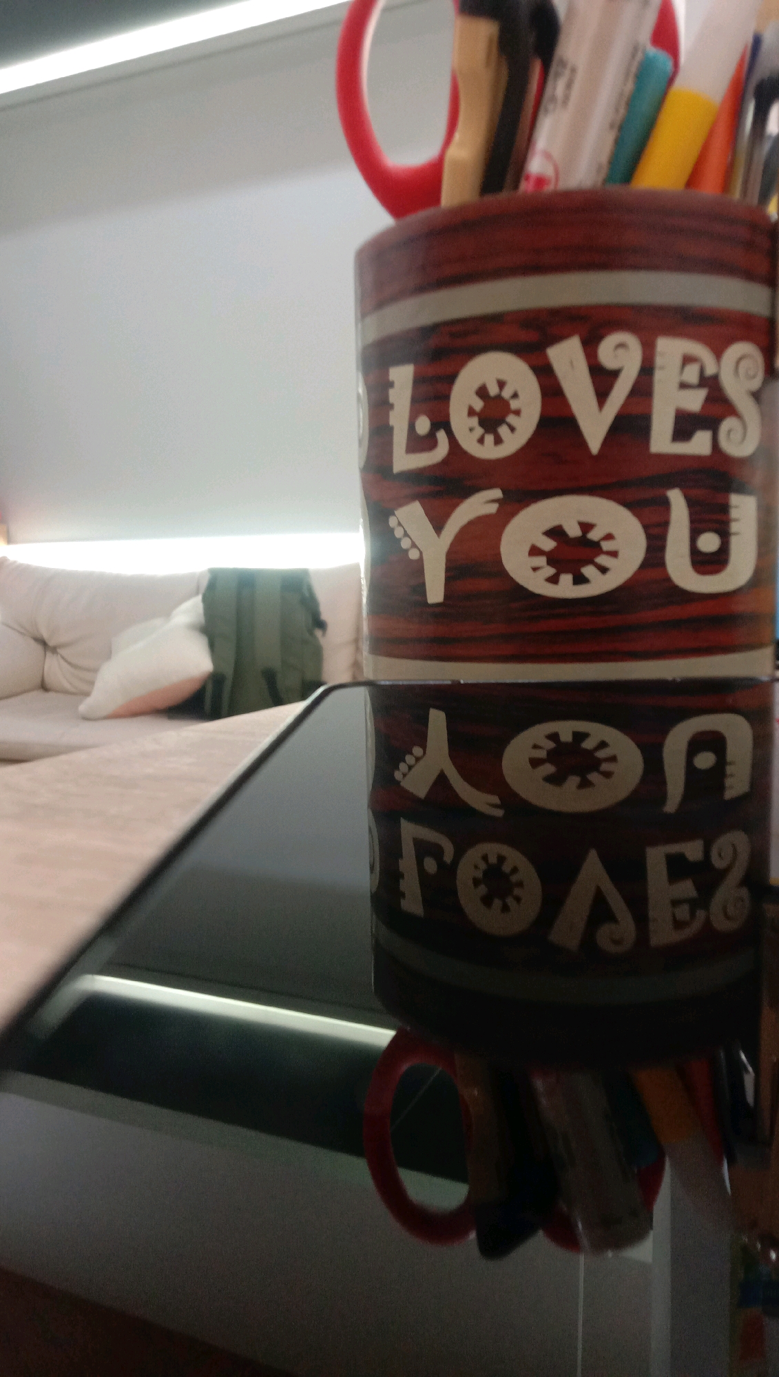 A close-up, slightly angled shot shows a red and black patterned mug filled with pens, pencils, and scissors. The mug has white text that reads "LOVES YOU" on the top half and appears to be a reflection of the same text on the bottom half. The mug is on a reflective, black surface, possibly a table, in front of a blurred background. The background shows a light-colored couch with pillows and a green backpack. A bright, horizontal light source is visible at the top of the frame. The scene is indoors, likely a living room or office. There are no people visible in the image.