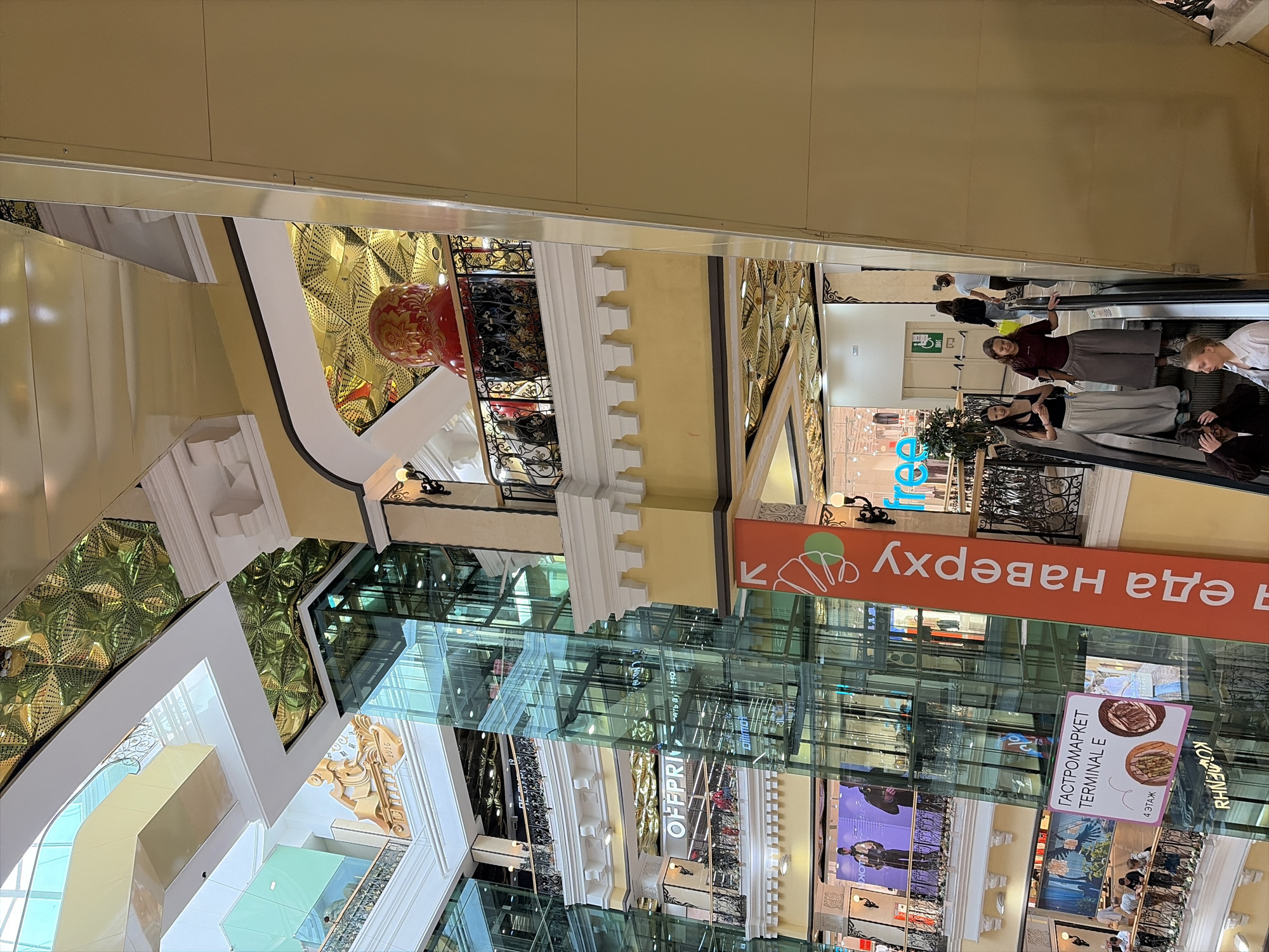 The image captures the interior of the "Greenwich" shopping mall in Yekaterinburg, Russia. The scene is a multi-level atrium featuring a large glass elevator shaft and escalators. On the right side of the frame, an escalator is visible with several individuals descending. In the foreground, two young women are on the escalator, one wearing a maroon top and gray pants, the other a black top and white pants. Further down, another young woman in a white shirt is visible. On the left side, the glass elevator shaft reveals multiple floors of the mall, with shops and people visible. One shop has a sign that reads "OFFPRICE." A large, red, matryoshka doll decoration is visible on an upper floor balcony, overlooking the atrium. The mall's interior design includes ornate ceilings with gold patterned details and yellow and white decorative elements on the walls and balconies. A prominent orange banner with white text, "Я еда наверху" (meaning "I am food upstairs"), is displayed vertically on the left side of the image, alongside arrow graphics. Signs for "ГАСТРОМАРКЕТ TERMINAL E" and "4 ЭТАЖ" (meaning "4th floor") are also visible on the lower left. The overall impression is of a bustling, modern shopping environment.