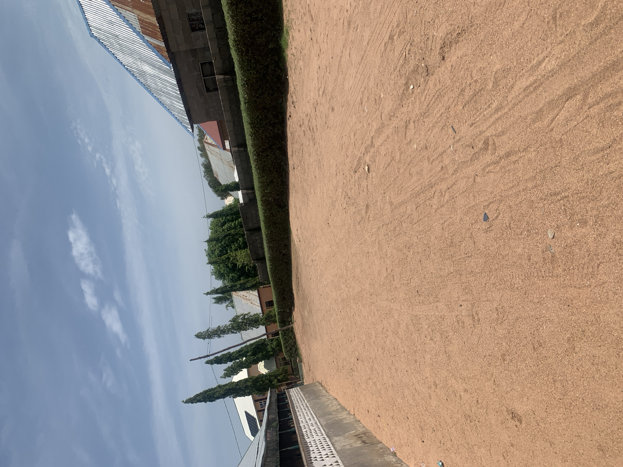 The image depicts a scene in Jauro Yinu, Nigeria, featuring a dirt path leading into the foreground. On the left side of the path, a low wall with a decorative white pattern runs along the edge of a building complex. Beyond this wall, solar panels are visible on a building's roof. The path is bordered by a well-maintained, dense green hedge. In the middle ground, several tall, slender trees stand out, interspersed with buildings exhibiting various roof types, including corrugated metal and what appears to be red roofing. The background is dominated by the sky, which is a mix of blue with scattered white clouds. The overall impression is of an outdoor area in a populated settlement.