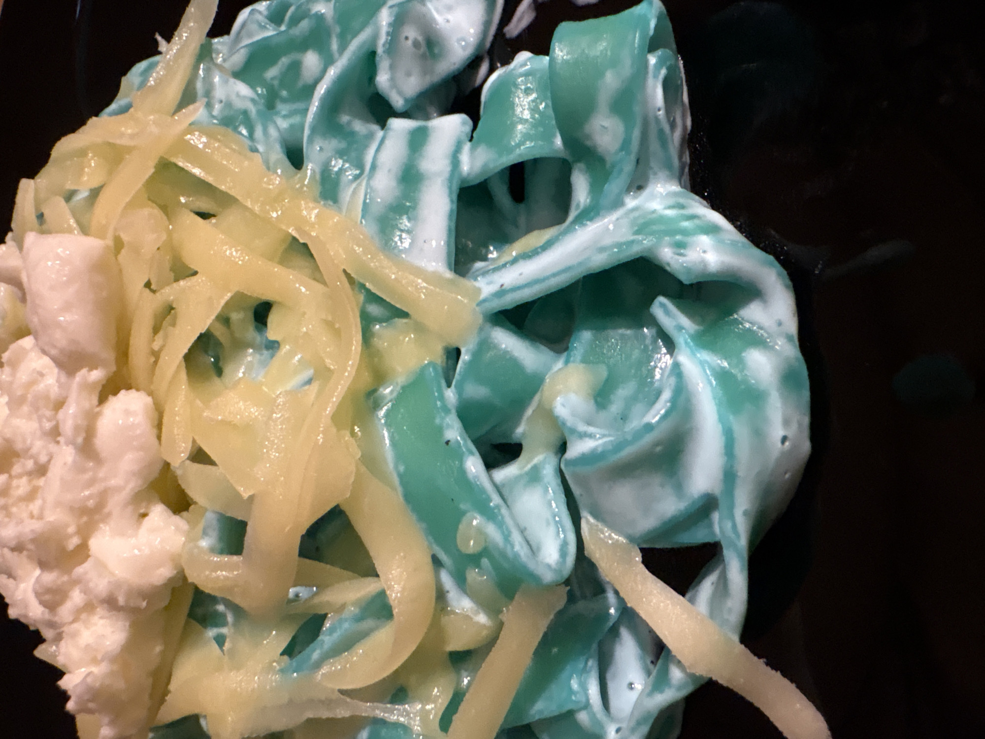 A close-up overhead shot displays a colorful dish resting on a dark, likely black, surface. The primary component is a portion of wide, flat pasta, possibly tagliatelle, which is distinctively colored a vibrant blue-green. This pasta is generously coated in a thick, creamy white sauce or dressing, giving it a glossy and somewhat marbled appearance.
Overlying a significant portion of the pasta is a heap of shredded, translucent yellow substance, which resembles grated cheese or finely julienned vegetables. To the far left of the image, there is a large cluster of a white, lumpy, and crumbly substance, suggestive of cottage cheese, ricotta, or a similar fresh dairy product.
The arrangement suggests a prepared meal, possibly pasta with a unique coloring and various toppings. There are no discernible activities or events taking place other than the presentation of the food. The image provides no visible text, and there are no specific cues within the frame to indicate the time of day, weather, or a particular geographical location. The focus is entirely on the textures and contrasting colors of the food.
