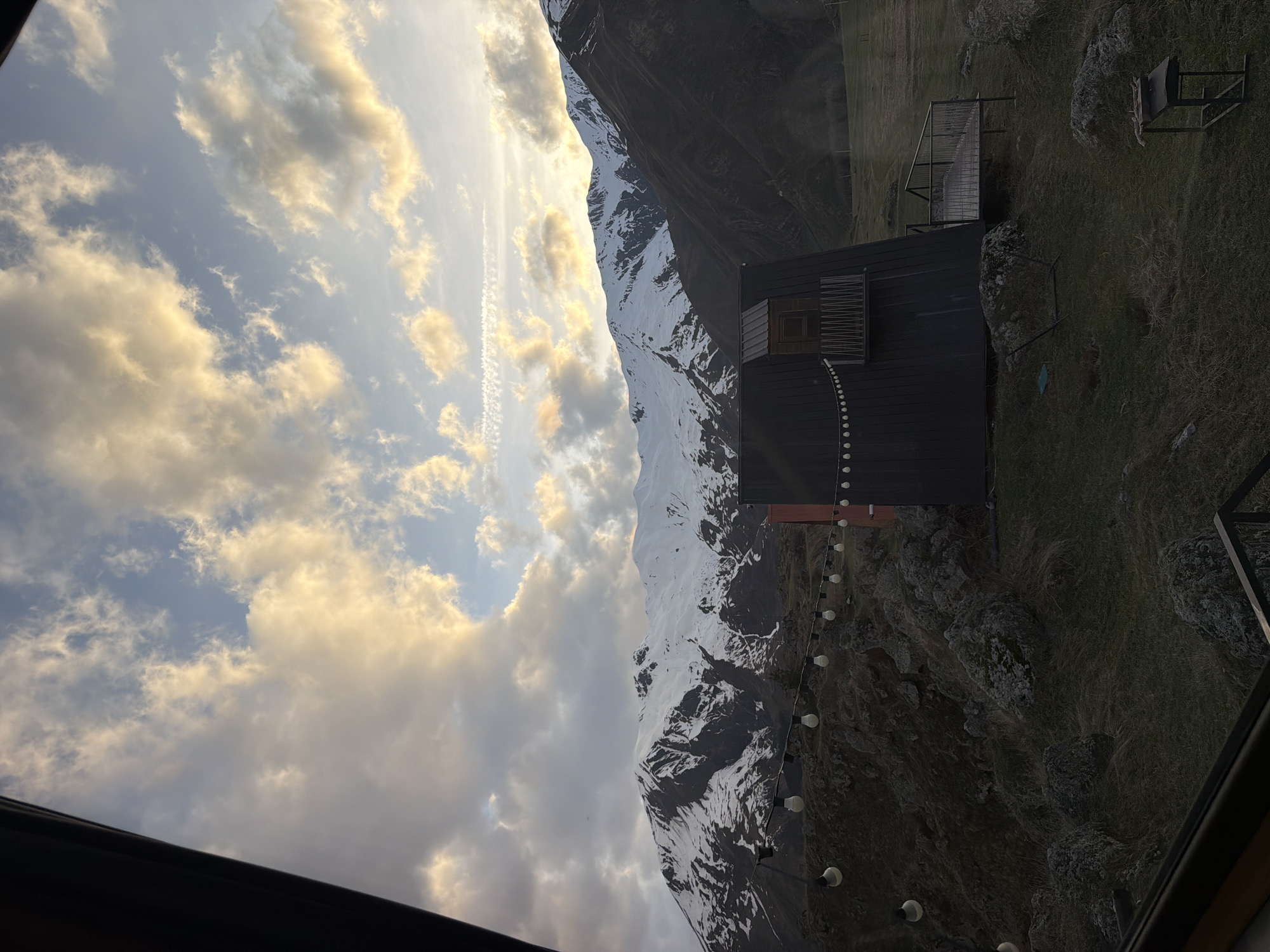 The image shows a landscape with a mountain range partially covered in snow, viewed from inside a building in Pkhelshe, Georgia. A dark-colored building with a wooden facade is situated on a grassy area next to a rocky cliff. A string of lights hangs along the edge of the building. The sky is filled with white and grey clouds, illuminated by sunlight. The view is partially framed by the interior structure of the building, visible in the foreground.