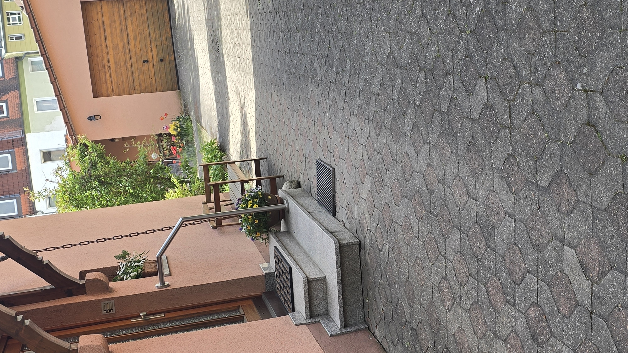 The image depicts a section of a streetscape in Zirndorf, Germany. The foreground features a peach-colored building with a visible door, window and wrought iron details including a chain, and flower boxes. In the background, there is a wooden garage door and neighboring buildings with multiple windows and varying colors of paint. The street itself is paved with hexagonal concrete pavers. A low stone retaining wall with a grated section runs alongside the sidewalk. There is a small wooden bench next to the wall, with flower pots on both sides.