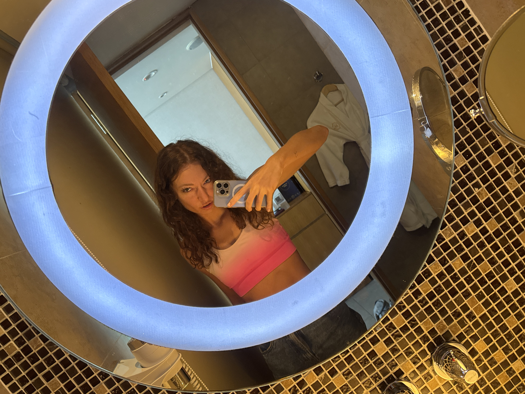 A young woman with long, curly hair is taking a selfie in a bathroom mirror with a circular LED light. She is wearing a pink and white gradient sports bra and denim jeans. She is holding a smartphone in her right hand. The mirror is surrounded by a mosaic tile pattern. In the mirror's reflection, a white bathrobe hangs on a hook, and there are glimpses of the bathroom interior. The faucet has a Kohler logo on it, and the environment appears to be in a hotel bathroom, as bathroom amenities are on the counter. Đà Nẵng, Vietnam.