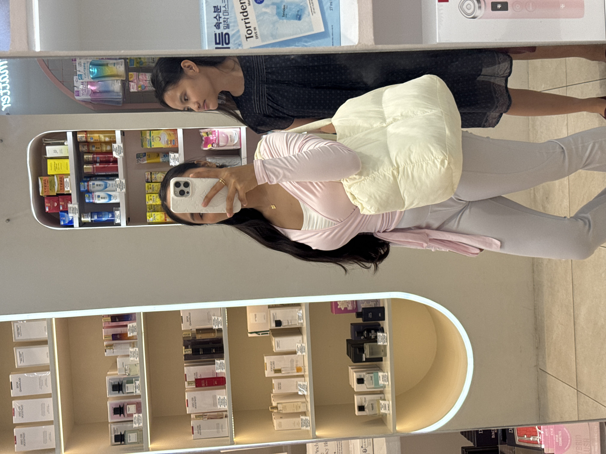 The image is a reflection in a mirror of two women in what appears to be a retail setting. The younger woman is wearing light-colored pants, a light pink wrap top, and a white shoulder bag. She is holding a phone and appears to be taking a picture of herself in the mirror. The other woman is standing to the right of the younger woman and is wearing a dark, patterned dress.
The background features shelves stocked with various products, including skincare items and perfumes. The product labels suggest they are from Korean brands. Also the OCR labels suggest it could be located in the city of Davao City, Philippines.