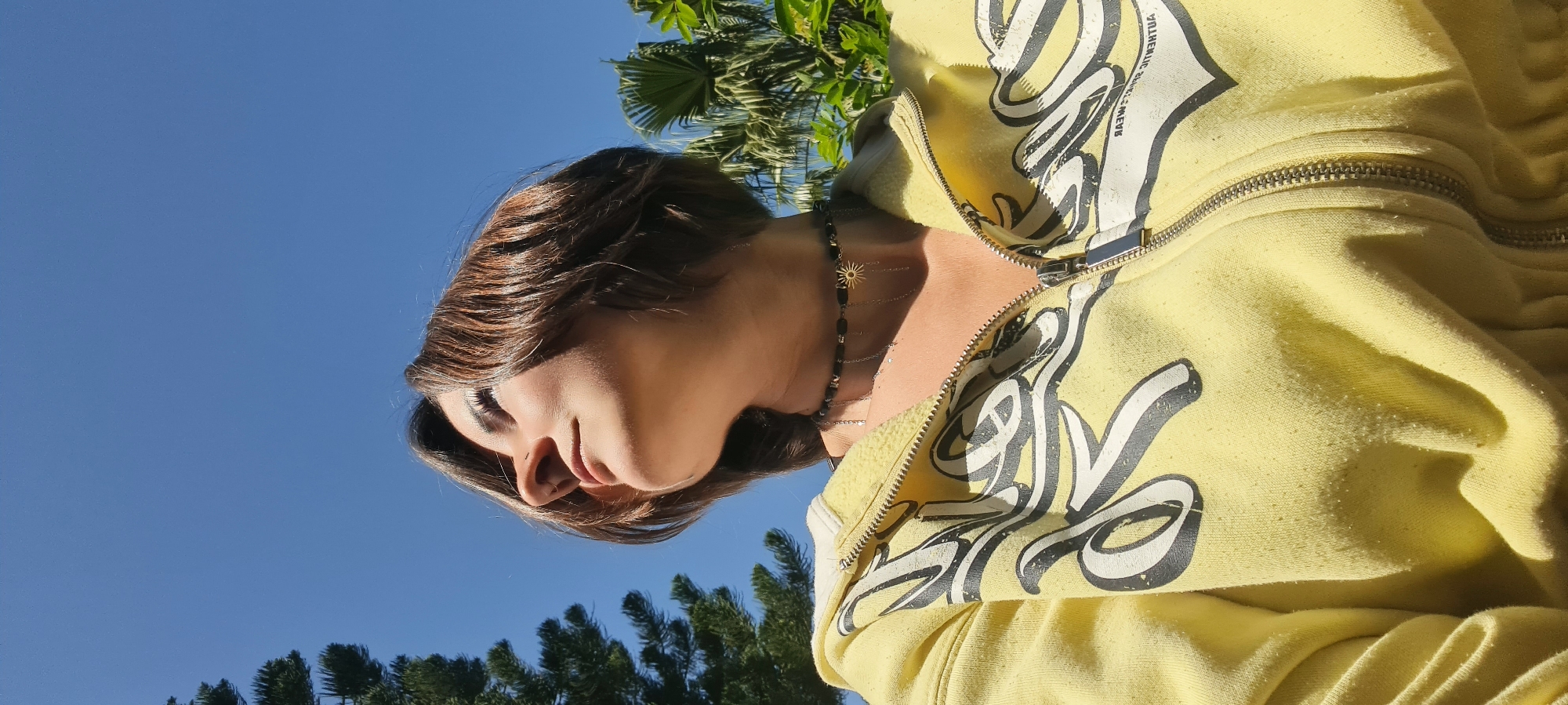A low-angle, medium shot depicts a person wearing a yellow zip-up hoodie with a black and white graphic print. The hoodie's zipper is partially open, revealing a necklace consisting of black beads and a gold-colored sun pendant. The person has short, brown hair and a light complexion. The backdrop includes a clear blue sky and green foliage, suggesting an outdoor location. The presence of palm trees implies a warm climate. The location is Nahariyya, Israel.