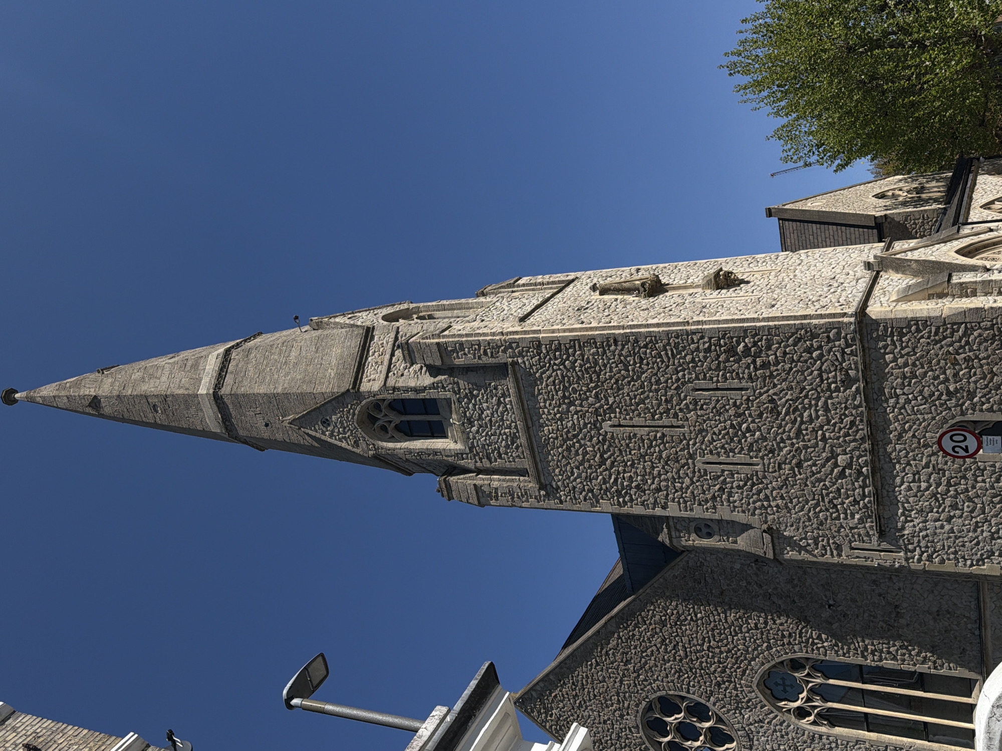 The image shows a tall church tower with a steeple, constructed from gray stone. The tower features arched windows and decorative stone elements. A speed limit sign of '20' is visible on the side of the tower. A tree is seen to the right of the tower. The sky is a clear blue. A lamp post and other street-level structures are also visible. The church is likely located in London, United Kingdom.