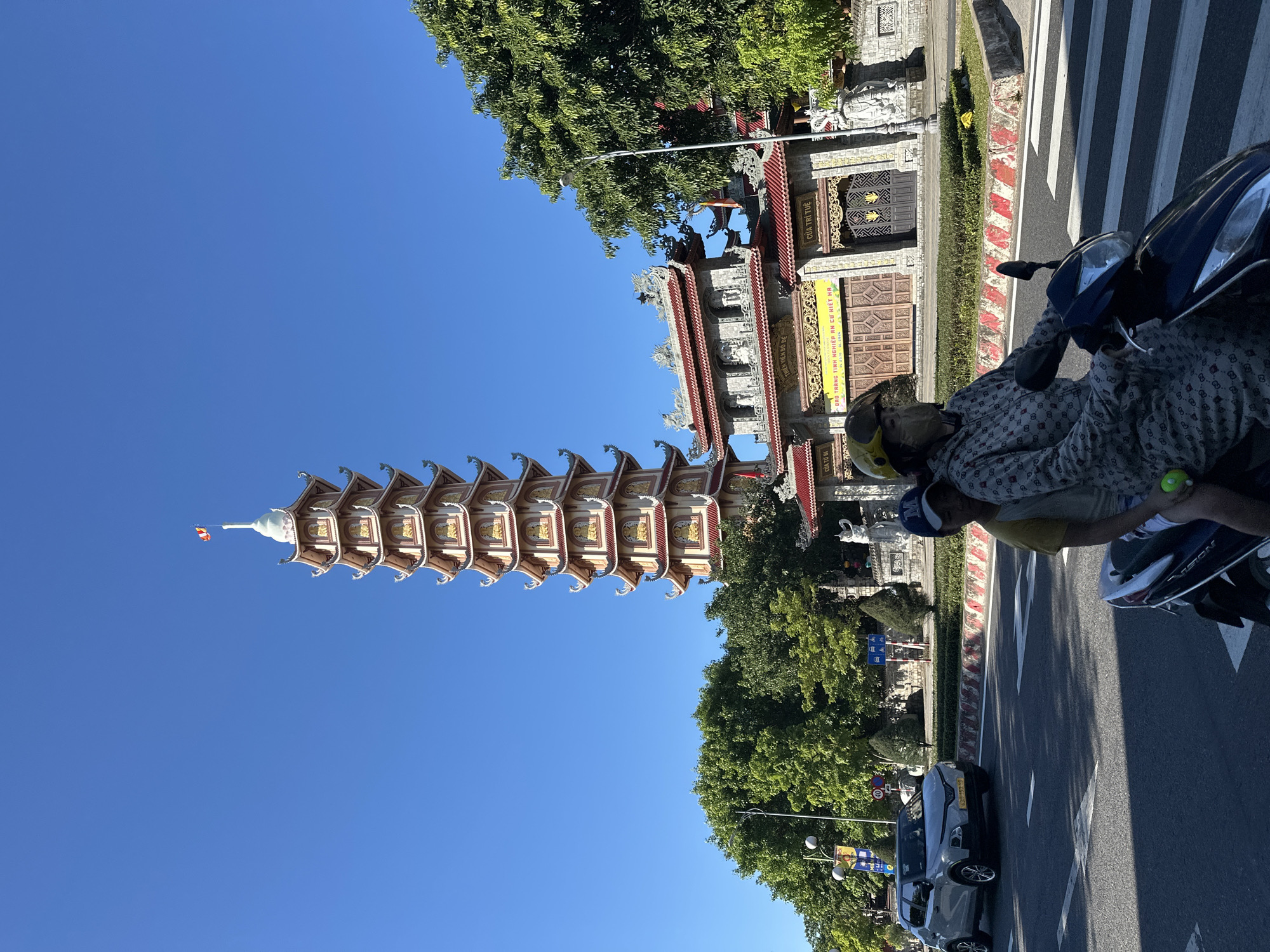 A tall, multi-tiered pagoda stands prominently against a clear blue sky in Đà Nẵng, Vietnam. The pagoda is elaborately decorated with ornate roofs and golden accents, featuring small Buddha statues or figures on each level. In front of the pagoda is a traditional temple complex with a red-tiled roof and decorative stonework. A street runs in the foreground, where a light-colored car is driving, and a person is riding a scooter. The scooter rider is wearing a patterned long-sleeved shirt, a yellow helmet, and a face mask. Lush green trees frame the temple and pagoda, and streetlights are visible. The scene captures a sunny day with clear visibility.