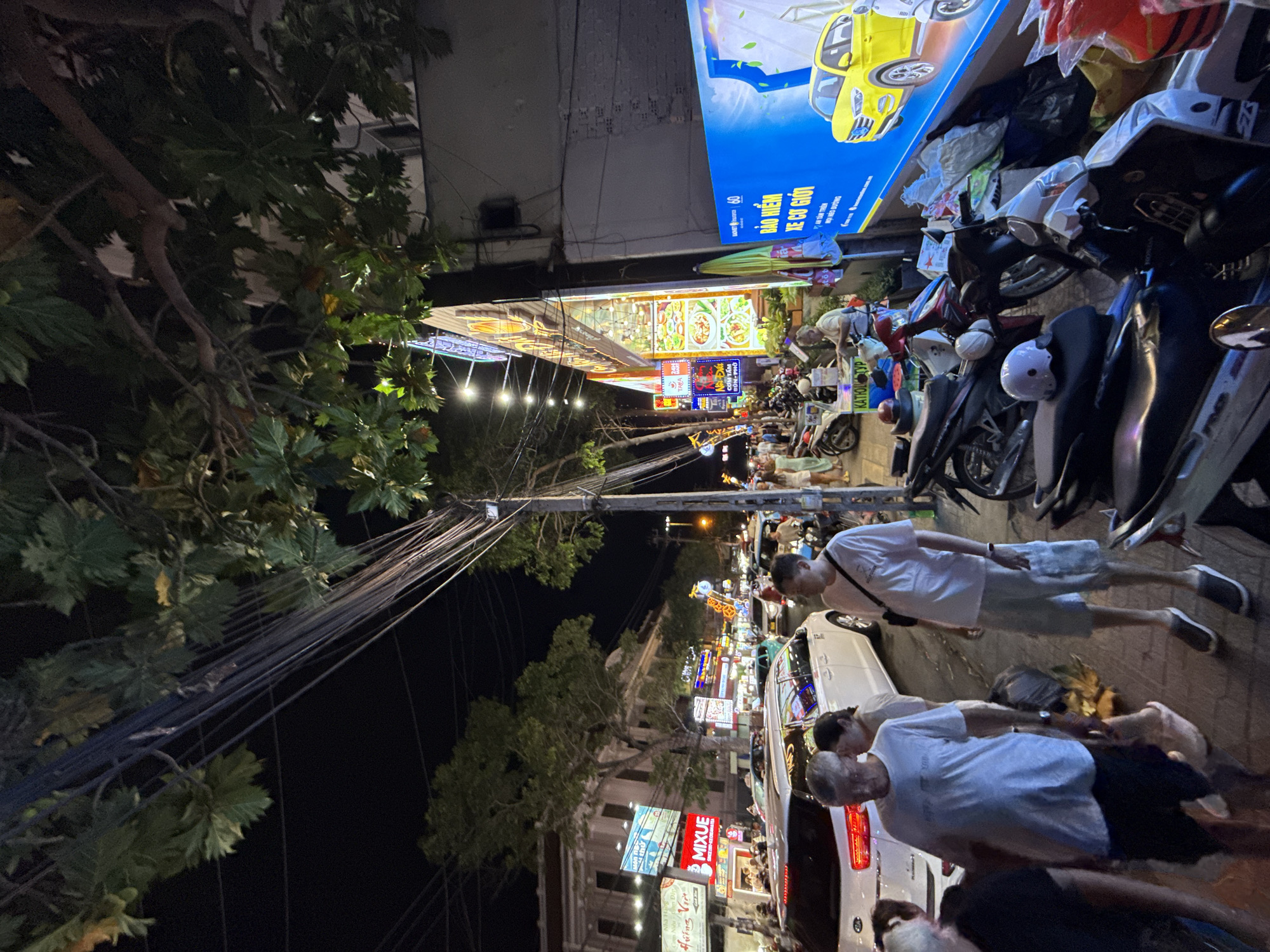 The image depicts a bustling street scene at night in Nha Trang, Vietnam, likely a commercial or entertainment district. A wide variety of shops and businesses line the street, illuminated by bright, colorful signs and lights. These signs feature text in Vietnamese and some English, advertising products and services, including food, telecommunications, and insurance.
The street is heavily trafficked with numerous motorcycles, cars, and pedestrians. People are walking along the sidewalk and crossing the street, some in groups and others individually. Many of the motorcycles are parked on the side of the road. The atmosphere appears lively and dynamic, with a mix of commercial activity and pedestrian movement.
The environment is characterized by overhead electrical wires, trees, and buildings. The night sky is dark, with artificial lighting from the shops and streetlights providing illumination. The presence of various signs, the types of vehicles, and the architectural style of the buildings suggest a Southeast Asian urban setting.
Specific details that can be observed include:
* A white Kia Sorento parked on the street.
* Several people, including men and women, dressed in casual attire.
* A large blue billboard on the right side of the image with text and an image of a yellow car, likely an advertisement for car insurance.
* A stall or food cart with an awning and a person operating it.
* A general sense of congestion and activity on the street.
The overall impression is of a vibrant and active urban environment at night.