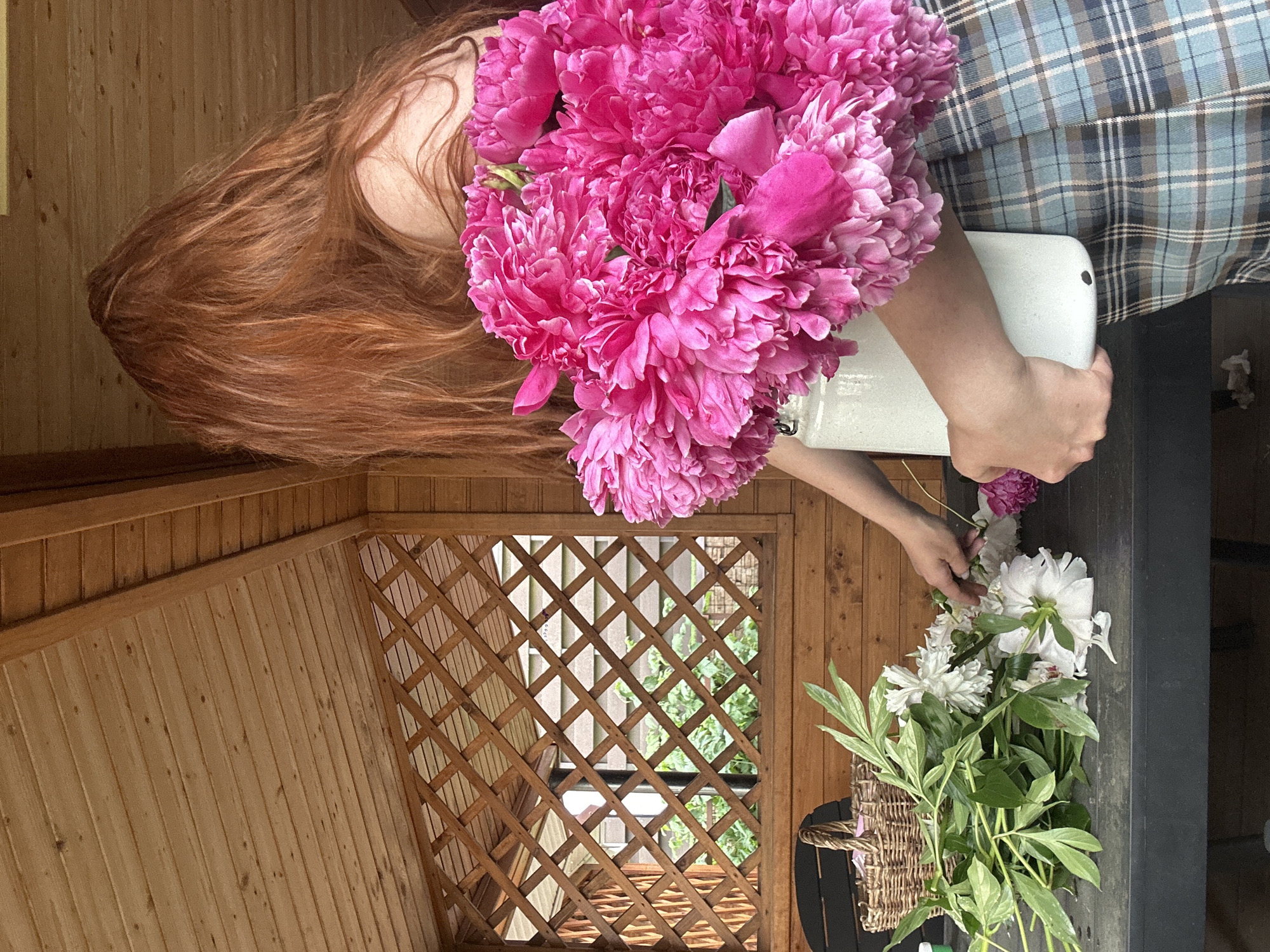A person with long, red hair is standing in a wooden gazebo, holding a large bouquet of bright pink peonies in a white ceramic vase. The person is wearing a blue and grey plaid skirt. In front of them, on a dark table, is a basket and a collection of white and green flowers, which the person appears to be arranging. The gazebo has a wooden slatted ceiling and a lattice-like wooden panel on the left side, revealing a green, leafy garden beyond. The overall scene suggests an outdoor, possibly domestic setting, within the city of Nizhny Novgorod, Russia.