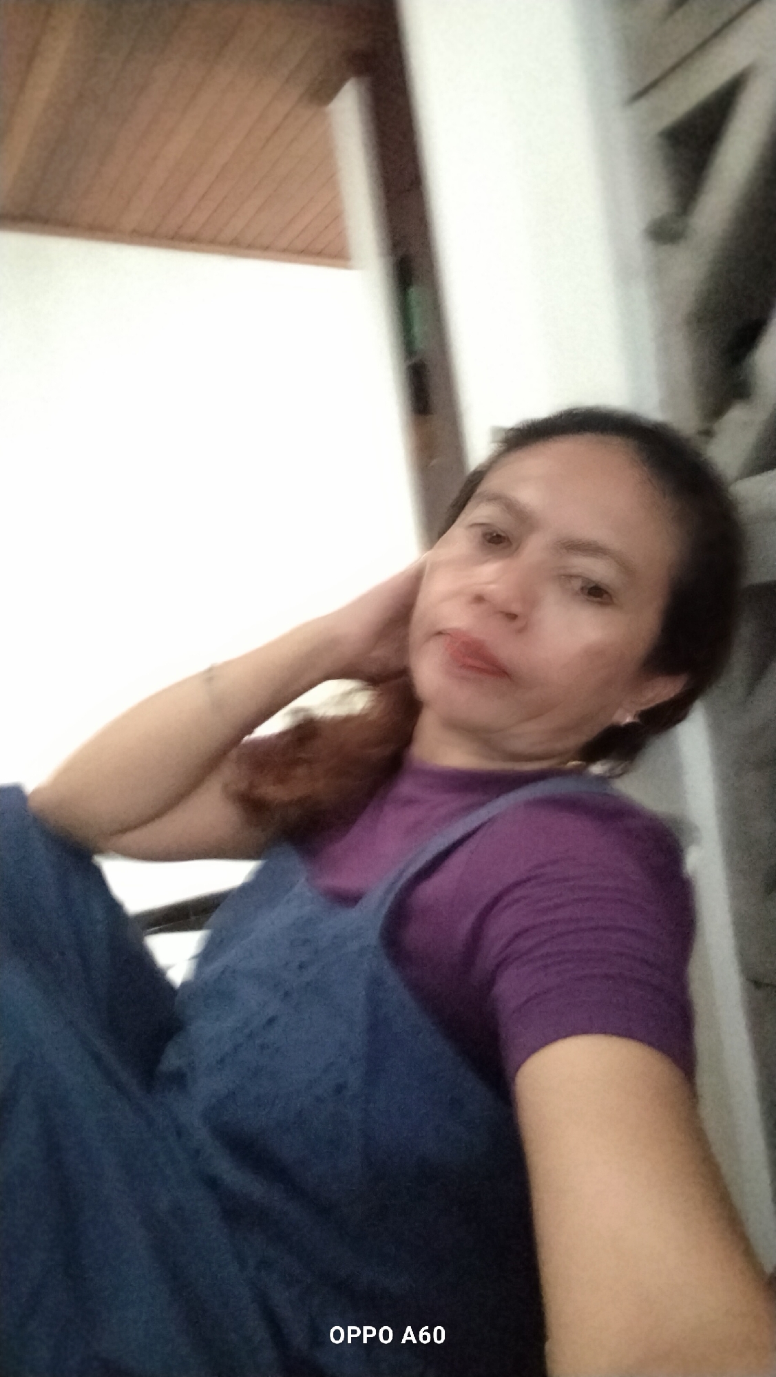 A woman, with dark hair and light brown skin, is reclining indoors, likely in her home in North Kuta, Indonesia. She wears a purple t-shirt and dark blue overalls. Her left hand is resting on the back of her head, and her right arm is extended downwards. The background features a white wall and a wooden ceiling. The image is a selfie taken with an OPPO A60 phone.