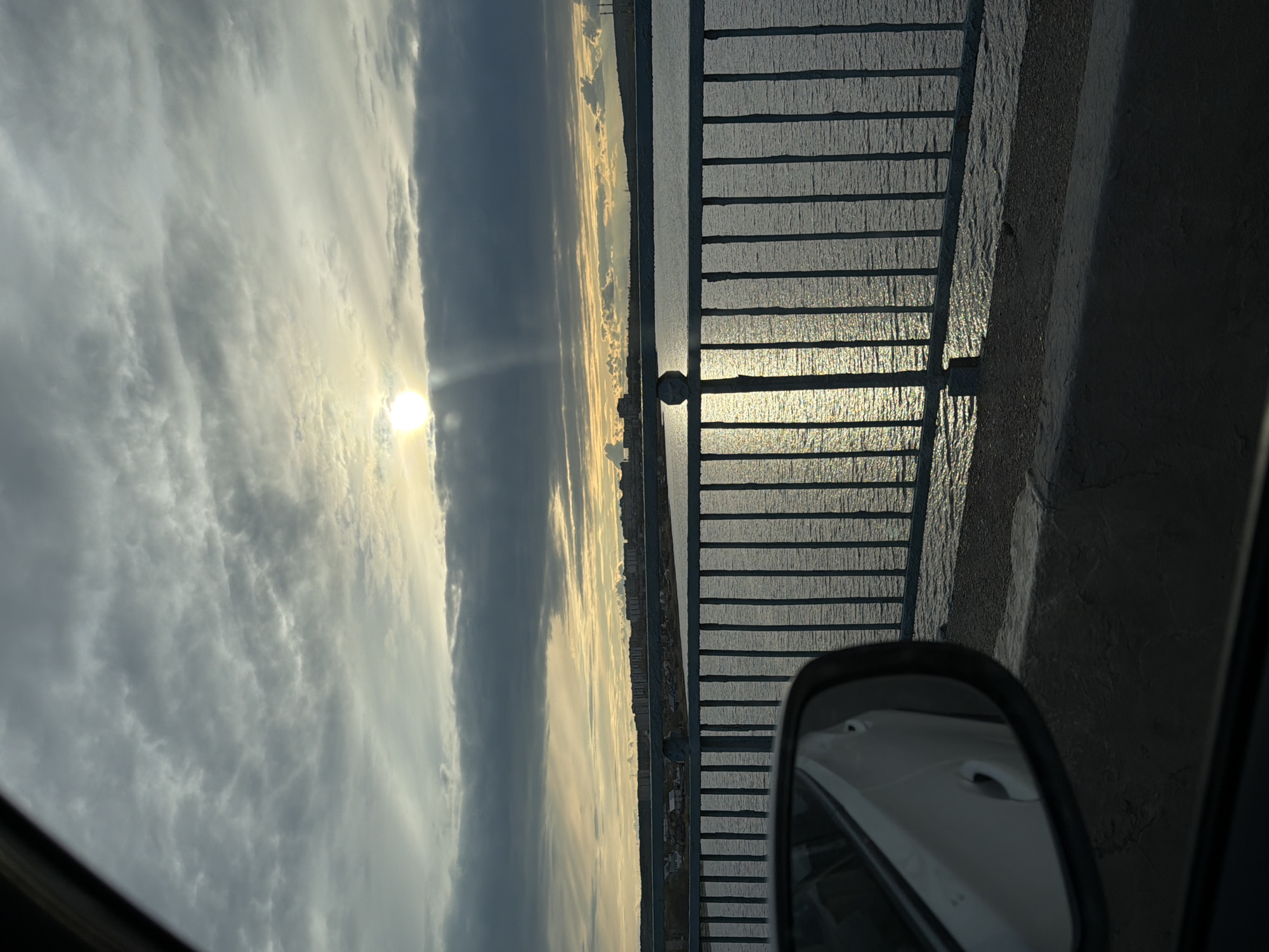 The image depicts a serene waterscape captured during either sunrise or sunset, viewed from the perspective of a vehicle. In the bottom right foreground, the side mirror of a car is visible, reflecting part of the car's white body.
A dark metal fence or railing stretches across the right side of the frame, running horizontally and bordering a large expanse of water. The water's surface shimmers with bright, golden reflections from the sun, indicating the "golden hour." The sun, a brilliant disc, is positioned on the left-center, partially obscured by clouds but still radiating intense light.
The sky is filled with sweeping, elongated clouds in various shades of gray and white, some illuminated by the golden sunlight. Along the distant horizon line across the water, a subtle landscape featuring what appears to be buildings or a cityscape is visible. The overall ambiance is peaceful and scenic, highlighting the interplay of light, water, and clouds. There are no people visible. Given the location context of Kamyshin, Russia, this large body of water is likely the Volga River, and the scene is possibly observed from a bridge or embankment. No text is visible in the image.