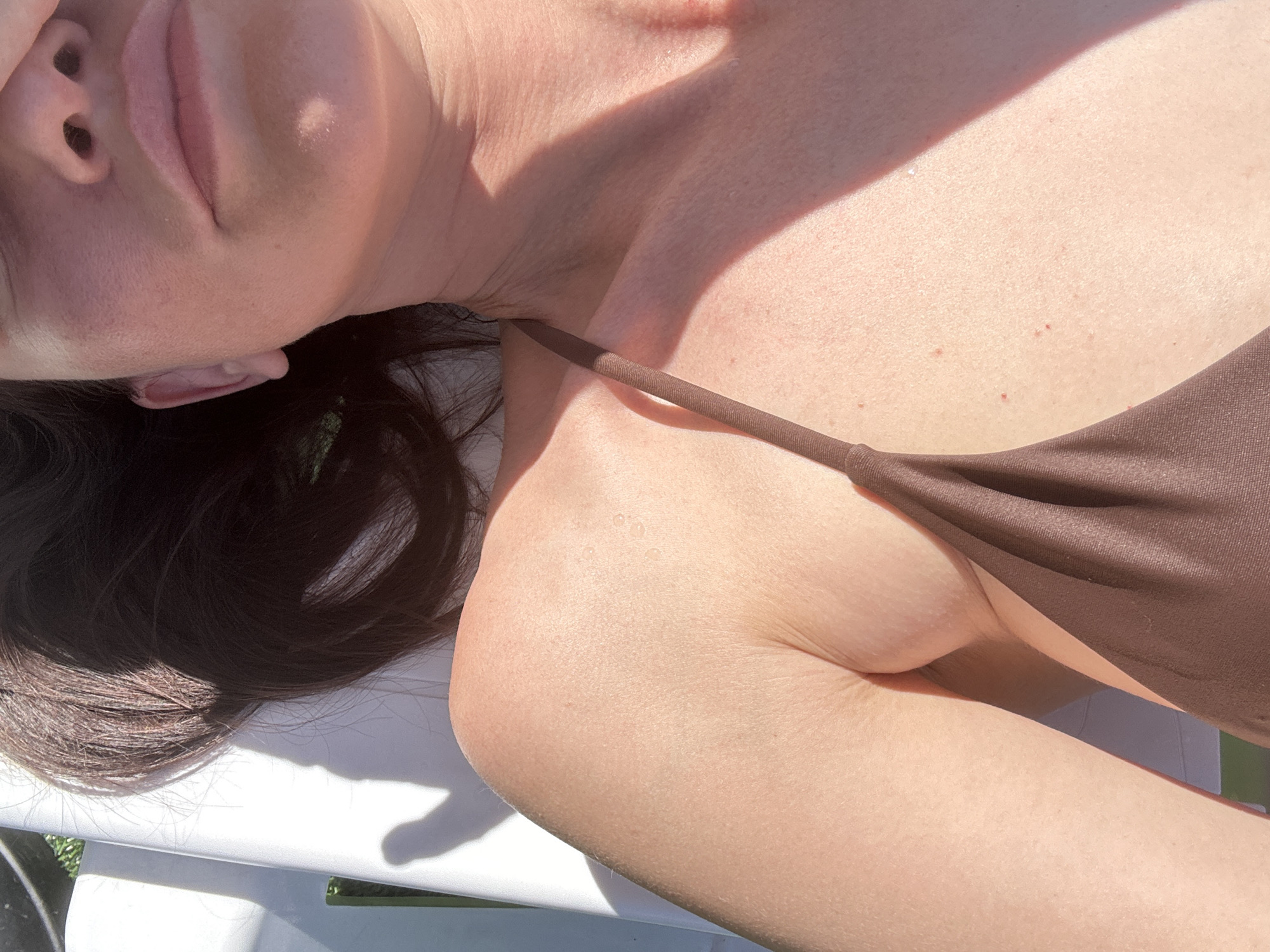The image shows a close-up of a person's upper body, lying on a white lounge chair outdoors. The person has dark hair and is wearing a brown bikini top. Sunlight casts shadows across their skin, highlighting a slight sheen of sweat or water droplets on their shoulder and chest. The background is slightly out of focus, showing hints of green foliage and what appears to be part of the lounge chair. The scene suggests a warm, sunny day, likely in a recreational setting.