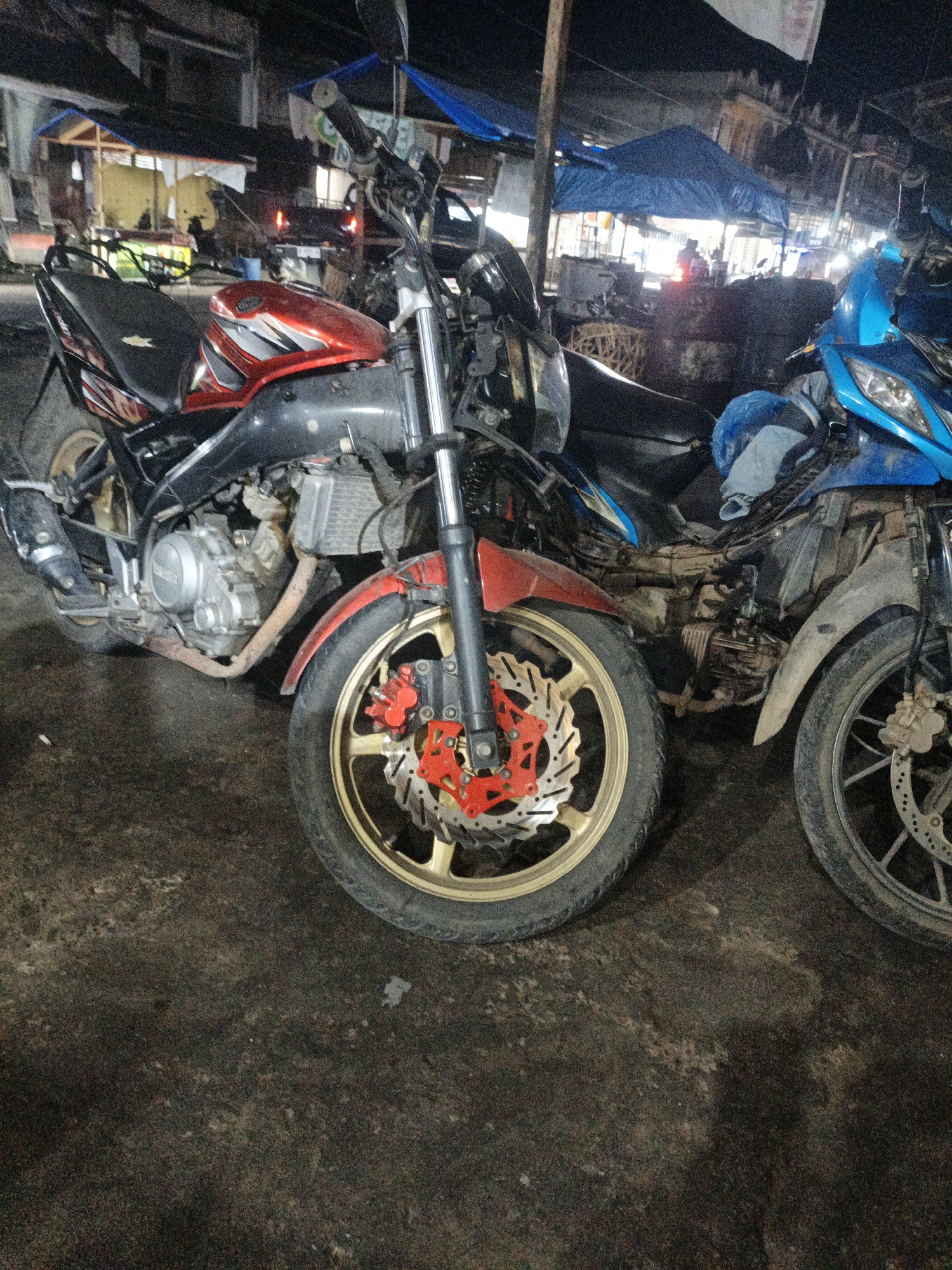 The image shows two motorcycles parked on a dark, presumably paved, surface. The motorcycle in the foreground is red and black, with a visible engine and gold-colored rims. The front wheel features a disc brake with red calipers. Behind it, to the right, is a blue motorcycle, also with a visible engine and a disc brake. The background is dimly lit and appears to be an outdoor area, possibly a market or roadside stall, with structures covered by blue tarps. The setting suggests it is nighttime or dusk. The location of Kruengmanyang, Indonesia, is not explicitly visible in the image itself but is provided as context for the scene. There are no people clearly visible in the image, though shadows or indistinct figures might be present in the background. The overall scene is gritty and appears to be in a public or semi-public space.