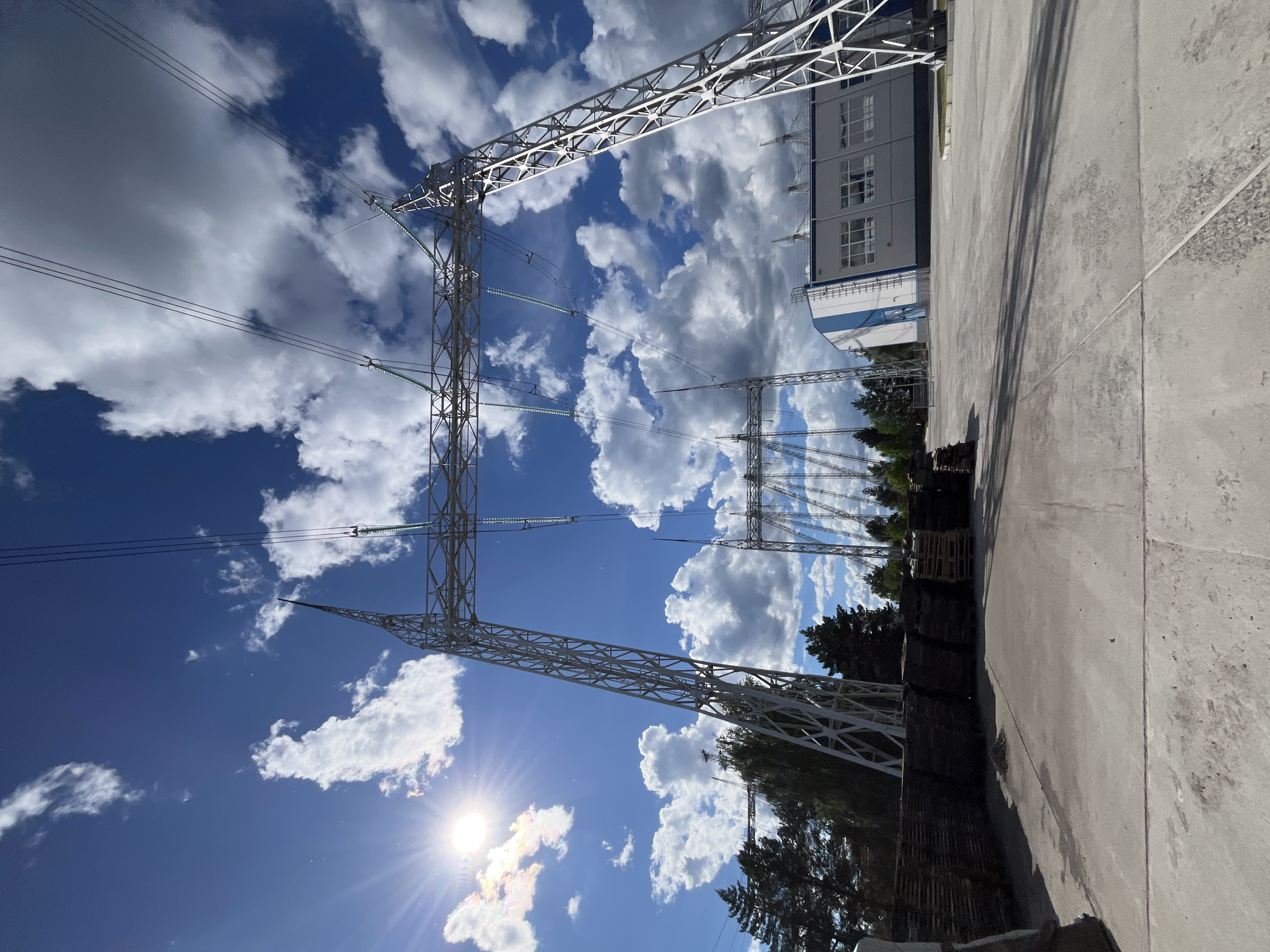 The image depicts a low-angle view of high-voltage power lines and pylons against a backdrop of a blue sky with scattered cumulus clouds. The sun is visible, shining brightly in the lower left portion of the frame. A building is visible in the background on the right side. Wooden pallets and vegetation are present along the ground. The location is the city of Chaikovsky, Russia.