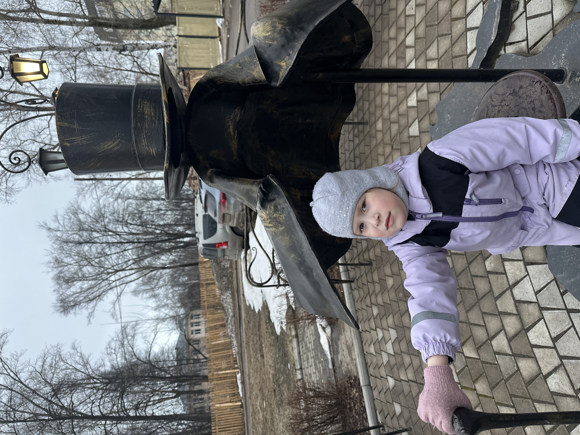 A child in a lavender and black jacket and a gray hat is sitting on a metal sculpture in the city of Chaikovsky, Russia. The sculpture appears to be a figure in a hat and coat. A lamppost with a light is connected to the sculpture. A white car is parked in the background, and bare trees are visible. The ground is paved with rectangular gray bricks, and there is a wooden fence in the distance. Patches of snow are present on the ground.