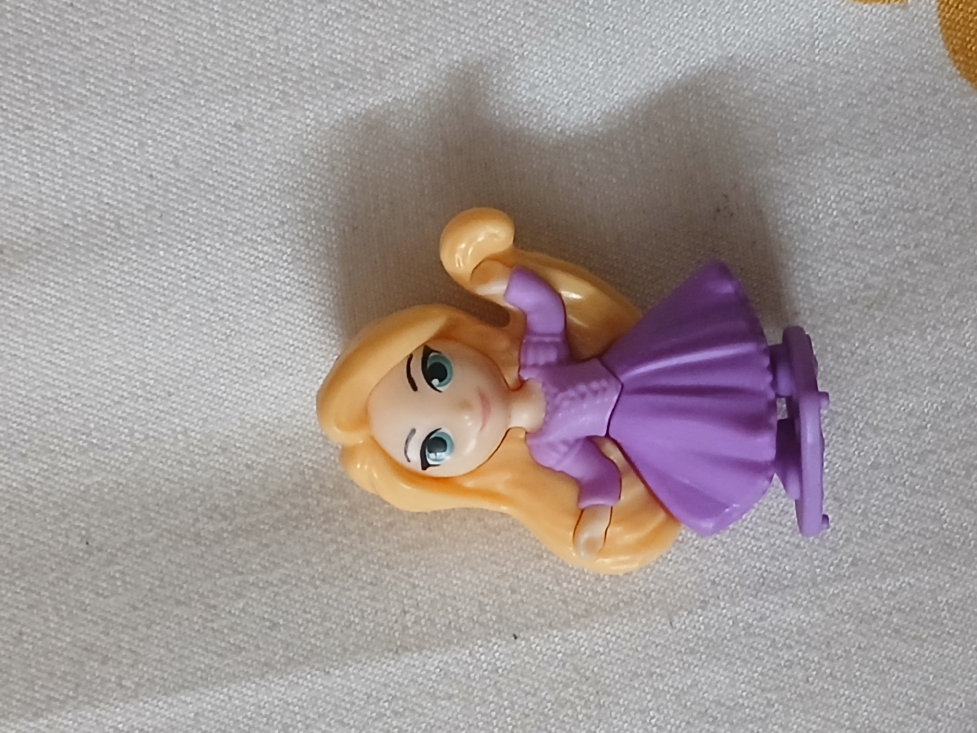 A small, plastic toy figurine of Rapunzel from Disney's "Tangled" lies on a textured, white surface, possibly fabric. The figurine is composed of light blonde hair, a light skin tone, and wears a lavender dress. The scene appears to be indoors with the lighting suggesting it's daytime. There are no visible signs that indicate the location is Bengaluru, India.