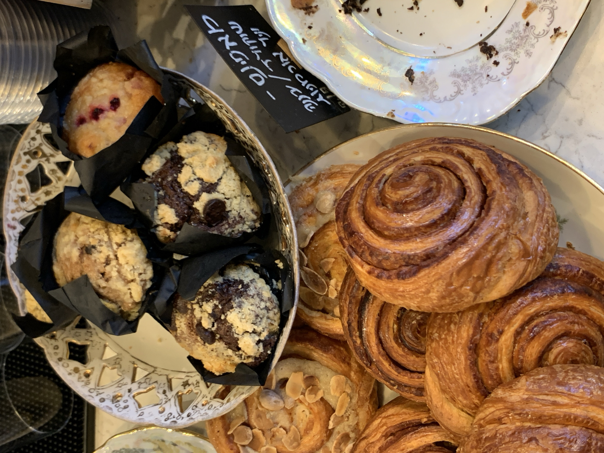 This is a close-up, overhead shot of a variety of baked goods, likely from a cafe or bakery. The subjects are primarily pastries: muffins, cinnamon rolls, and other swirled, layered baked goods. One muffin in the foreground appears to be a berry muffin with visible red berries. Another muffin is dark chocolate with a crumble topping. The cinnamon rolls are golden brown and appear flaky, with some topped with sliced almonds. The pastries are arranged on decorative platters, one of which is white with gold trim and has a cut-out pattern.
A black sign with white text is visible in the upper left quadrant of the image, angled towards the baked goods. The text is in Hebrew and appears to include prices or descriptions, with some English words like "NICCOLI" visible as well.
The lighting suggests an indoor setting, possibly during the daytime. The overall impression is one of a sweet treat selection, perhaps for breakfast or a coffee break. The focus is entirely on the food items, creating a visually appealing spread. The scene is clean and well-presented, indicative of a commercial establishment.
