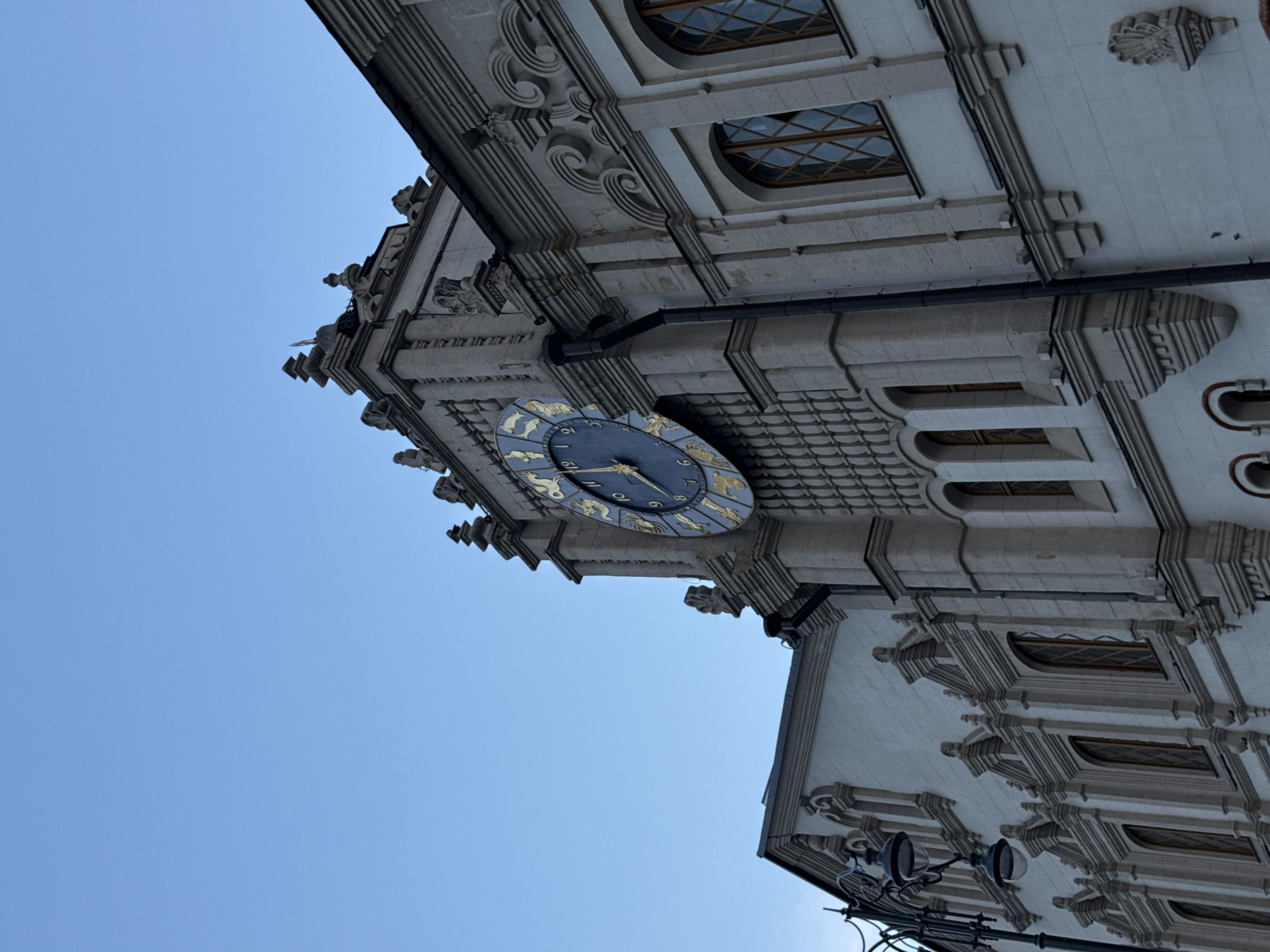 The image shows the ornate facade of a building with a large astronomical clock, set against a clear blue sky. The building is constructed from light-colored stone and features decorative elements such as cornices, window frames, and sculpted ornamentation. The clock has a dark blue face with golden zodiac signs marking the hours. The building's architectural style suggests a historic or monumental structure. The context of the city of Ukromne, Ukraine, suggests this could be a significant landmark or historical building within the town.
