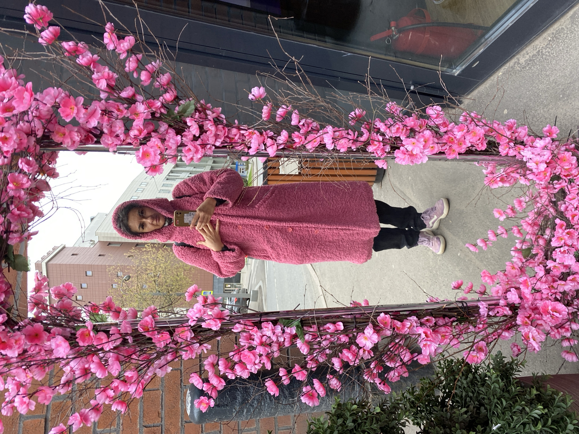 The image displays a young woman posing in front of a decorative arrangement of artificial pink cherry blossoms. The woman and the background are reflected in a large, vertical mirrored or highly reflective surface, which is framed by numerous branches covered in bright pink flowers.
The woman, with dark hair partially covered by a hood, is positioned centrally in the reflection, looking towards the camera with a neutral expression. She is wearing a long, hooded coat made of a textured, fuzzy pink material, black pants, and light purple sneakers with white soles. She holds a smartphone with a colorful, patterned case in her hands.
The reflected background indicates an urban setting, likely a street in Moscow, Russia. Multi-story buildings with various windows are visible, along with a street featuring a yellow road sign (possibly a pedestrian crossing) and distant traffic. The bright, overcast sky suggests daytime. In the immediate surroundings, parts of a brick wall and a dark green bush or plant are visible. Behind the reflective surface, a red fire extinguisher can be seen.
The scene appears to be a public or commercial space featuring a decorative floral installation, possibly designed for spring or a special event. The woman is engaging with the display, seemingly taking a photo or posing for one. No discernible text is clearly legible on any objects within the image.
