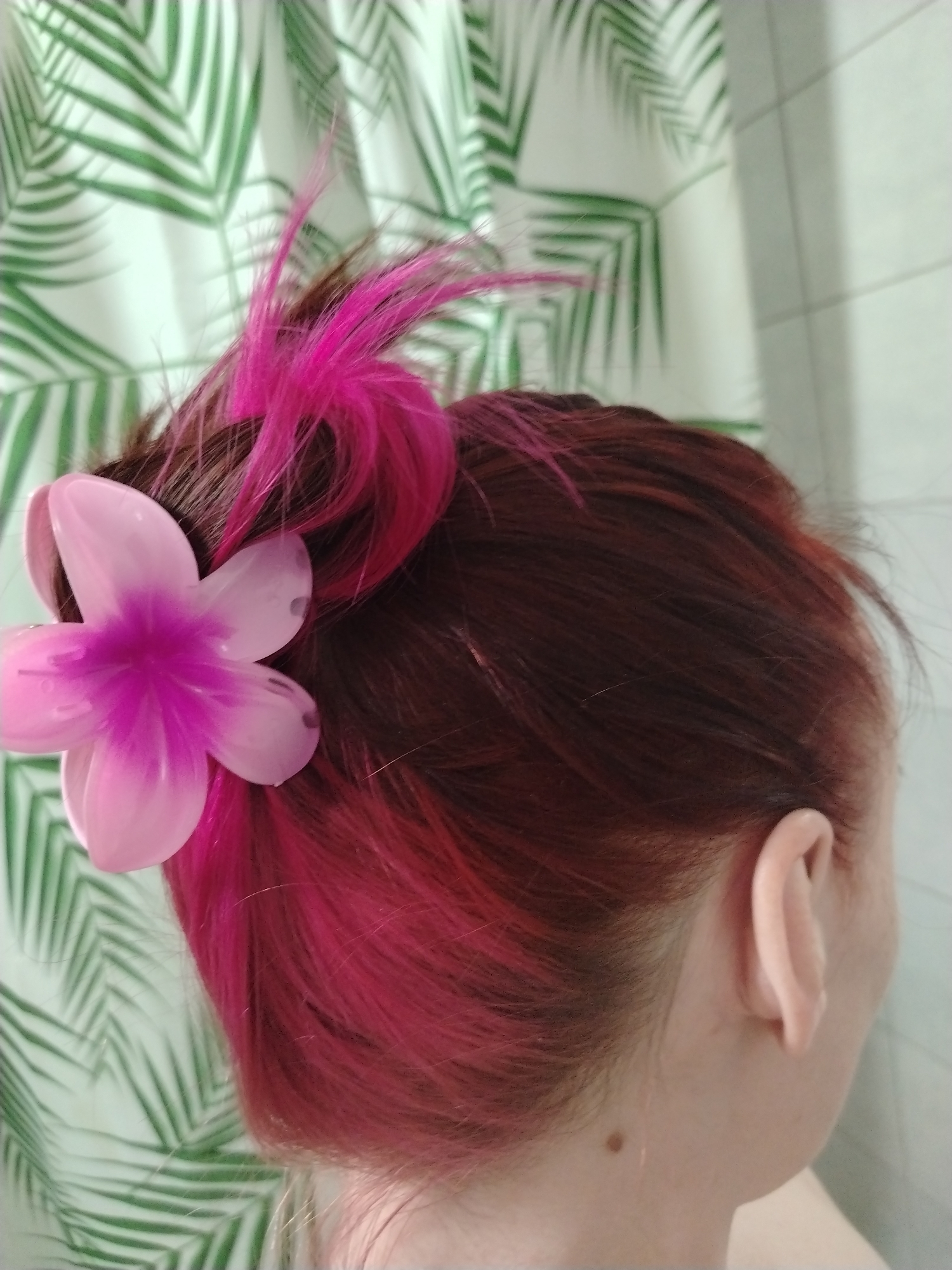 A close-up shot shows the back of a person's head, with auburn hair and vibrant pink highlights. The hair is styled in a bun, secured with a pink flower hair clip. The background features a shower curtain adorned with a pattern of large green tropical leaves. A small mole is visible on the person's neck. No other information is present in the image to suggest a location in Izhevsk or any specific city.