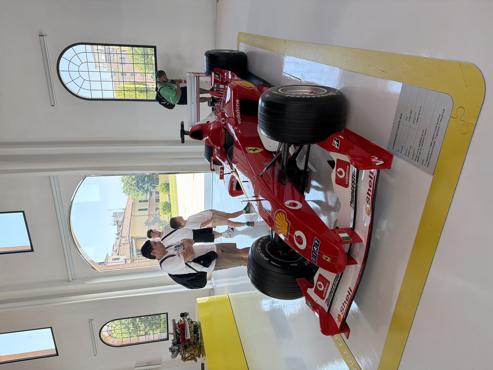 The image shows a red Ferrari F2003-GA Formula 1 car displayed indoors, likely in a museum or exhibition hall. The car is the central subject, positioned on a white floor with yellow demarcation lines. A descriptive plaque is placed in front of the car, detailing its specifications. Several people are present, observing the car. On the left, two men and a boy stand near a yellow display that holds a red engine. In the background, another person is seen looking at the car from behind. Large arched windows in the background offer a view of buildings and trees, suggesting an urban or semi-urban setting. The architecture of the building appears modern with high ceilings and white walls. Based on the car and the presence of a Ferrari engine, the location is very likely the Enzo Ferrari Museum in Modena, Italy.