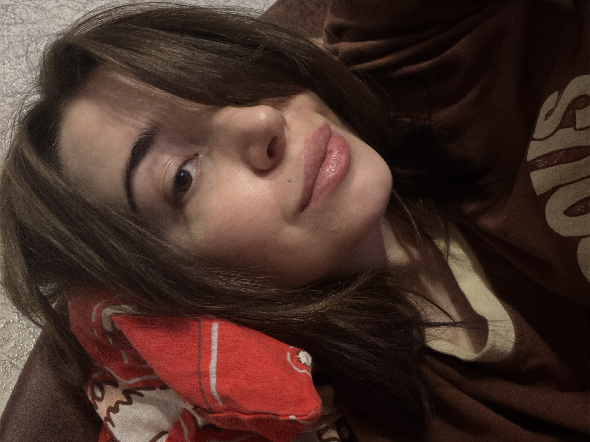 A close-up, head-and-shoulders shot shows a young Caucasian woman with medium brown hair and fair skin, looking toward the camera. She has a slight smile and her eyes are partially obscured by her hair. She is wearing a brown t-shirt with a lighter-colored crew neck. A red cloth with a white and black pattern is visible behind her left shoulder, tucked against her head. The background appears to be a textured wall. The lighting is dim, casting shadows.