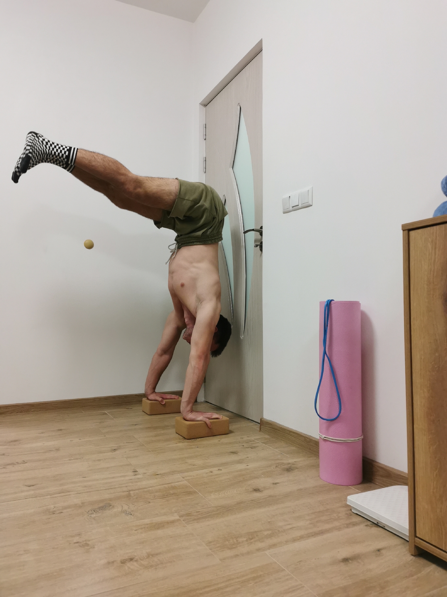 A man performs a handstand indoors against a white wall and door in what appears to be a home in Burgas, Bulgaria. He is shirtless, wearing olive green shorts and black and white checkered socks. His hands are placed on two light brown yoga blocks on a light wooden-look tiled floor, while his feet rest against a light-colored door with a translucent panel.
The man's body is inverted, forming an L-shape with his legs extended upwards and his head pointing towards the floor. A small, light-colored ball is stuck to the white wall on the left side of the frame, near the man's torso. On the right side of the image, a rolled-up pink yoga mat, secured with a blue resistance band, leans against the wall. A white digital weight scale is visible on the floor next to the yoga mat, partially obscured by a wooden cabinet. White light switches are mounted on the wall to the right of the door. The room is brightly lit, with clean white walls and a consistent flooring, suggesting a well-maintained interior. The man's expression is focused, typical for such an exercise.