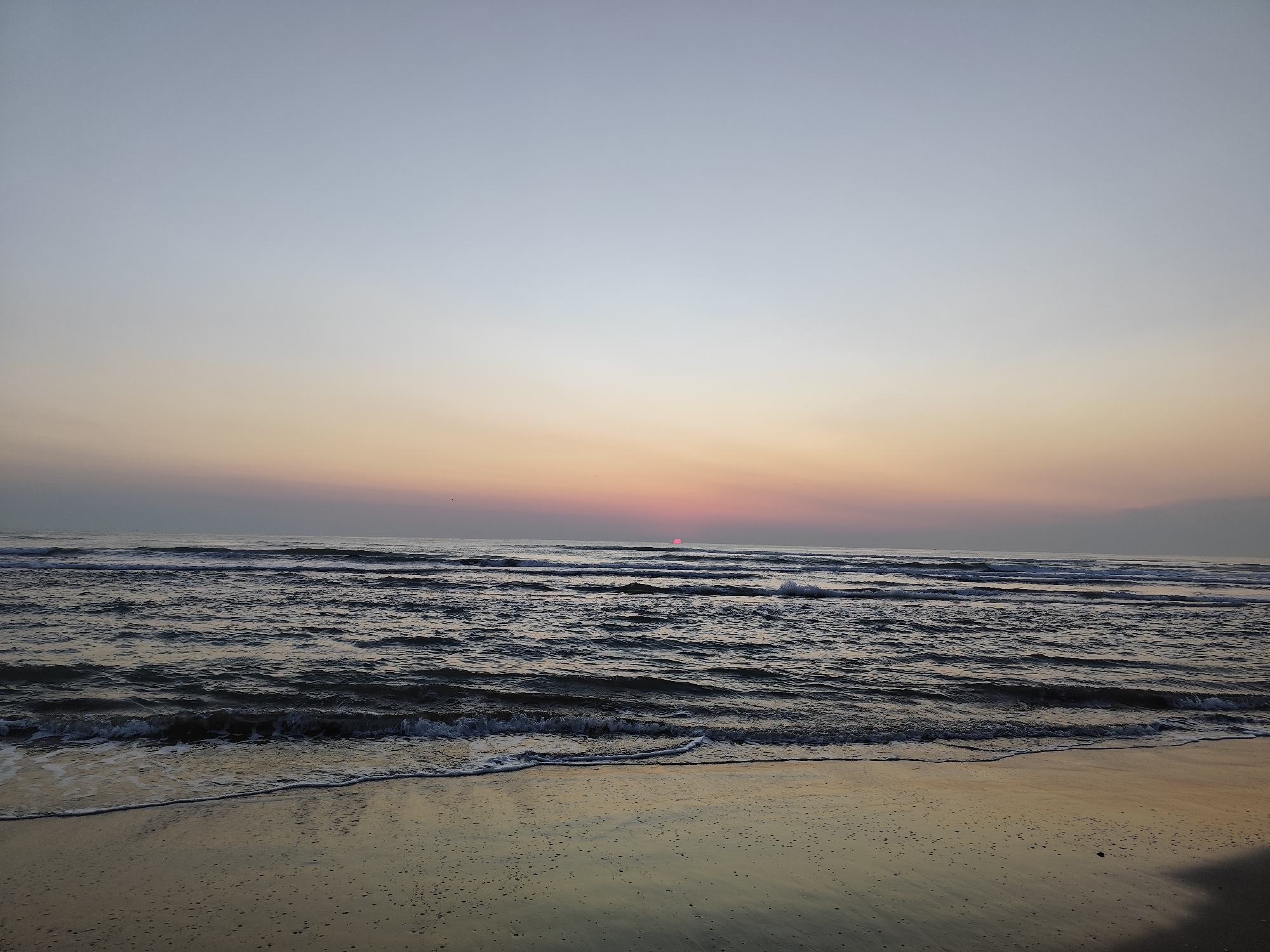 A wide shot captures a tranquil beach scene at dawn or dusk in Rimini, Italy. The sky transitions from a pale blue at the top to soft hues of pink and orange near the horizon, where a sliver of the sun is visible peeking above the ocean. Gentle waves roll onto the sandy shore, with a thin line of foam marking the water's edge. The wet sand reflects the muted colors of the sky. There are no people or man-made structures visible in this serene natural landscape.