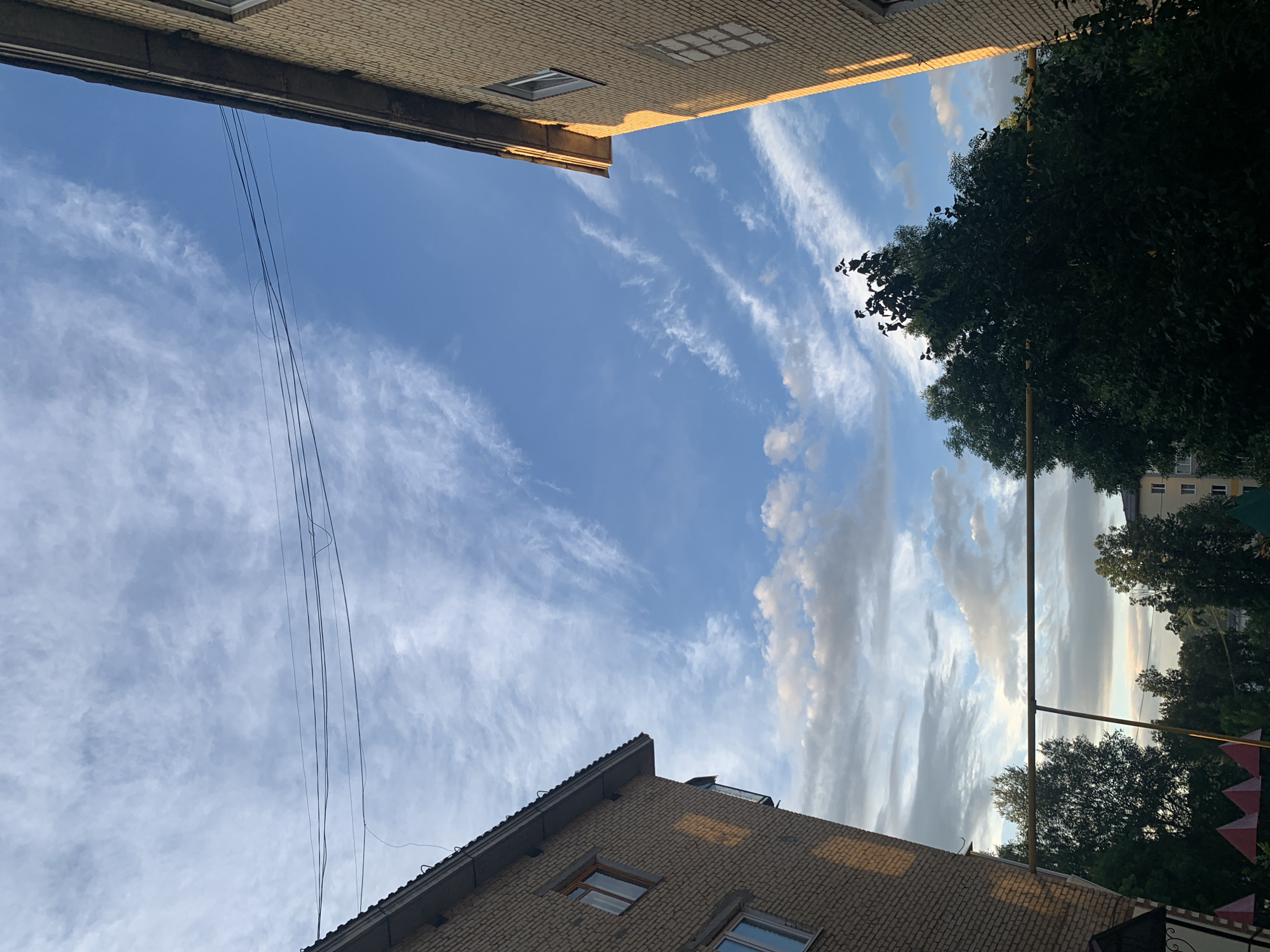 The image captures a view of the sky between two brick apartment buildings in Asha, Russia. The sky is a bright blue, with scattered cirrus clouds stretching across it. Power lines are visible against the sky, strung between the buildings. The buildings themselves are of a light brown brick construction, with visible windows. Green trees are present in the foreground and middle ground, with some foliage obscuring parts of the lower sections of the buildings. A metal frame, possibly part of a clothesline or playground equipment, is visible horizontally across the center of the image, behind the trees. The lighting suggests it is either early morning or late afternoon, with sunlight casting warm tones on the brickwork of the buildings. There are no people or animals visible in the image.