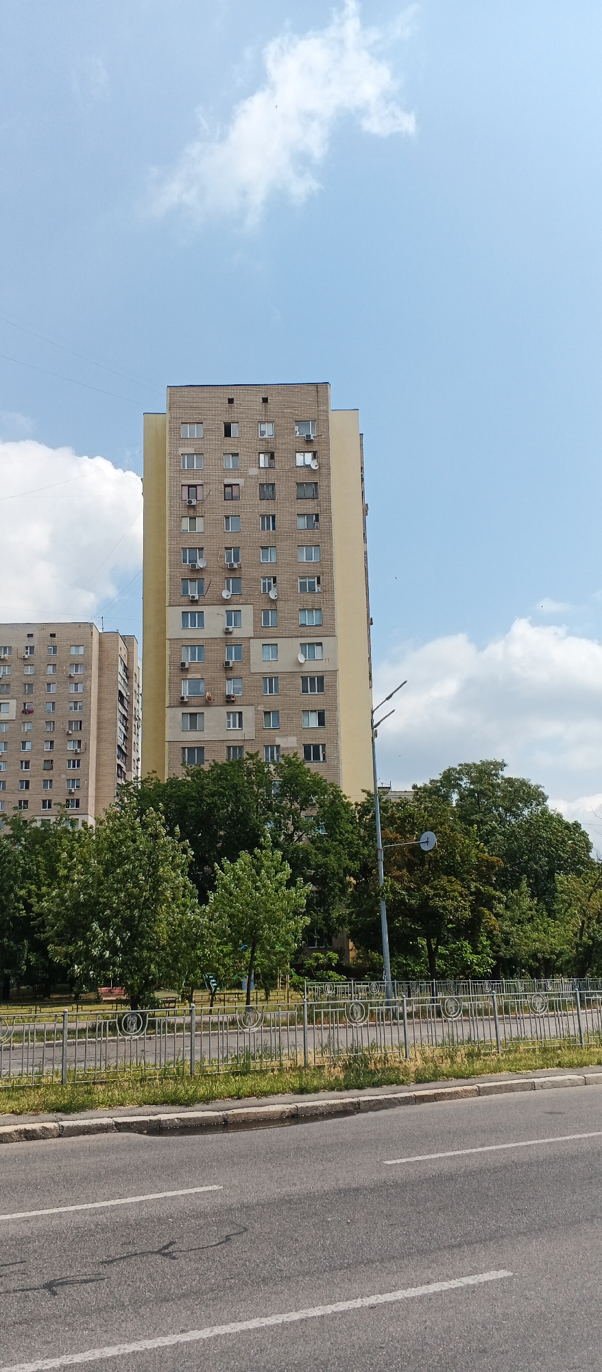 The image captures a street scene in Kyiv, Ukraine, on a clear day with scattered clouds. In the background, two tall apartment buildings dominate the skyline. The building on the right, painted in a pale yellow and beige brick pattern, has multiple windows and air conditioning units visible. To its left, a similar, but slightly shorter, apartment building with a grey brick facade is partially visible.
In the foreground, a paved road with white lane markings stretches across the lower portion of the image. Adjacent to the road is a sidewalk and a decorative metal fence. Beyond the fence, lush green trees form a dense line, partially obscuring a park-like area with benches and playground equipment. A street lamp with two fixtures stands near the fence, silhouetted against the sky. The overall impression is a typical urban residential environment.