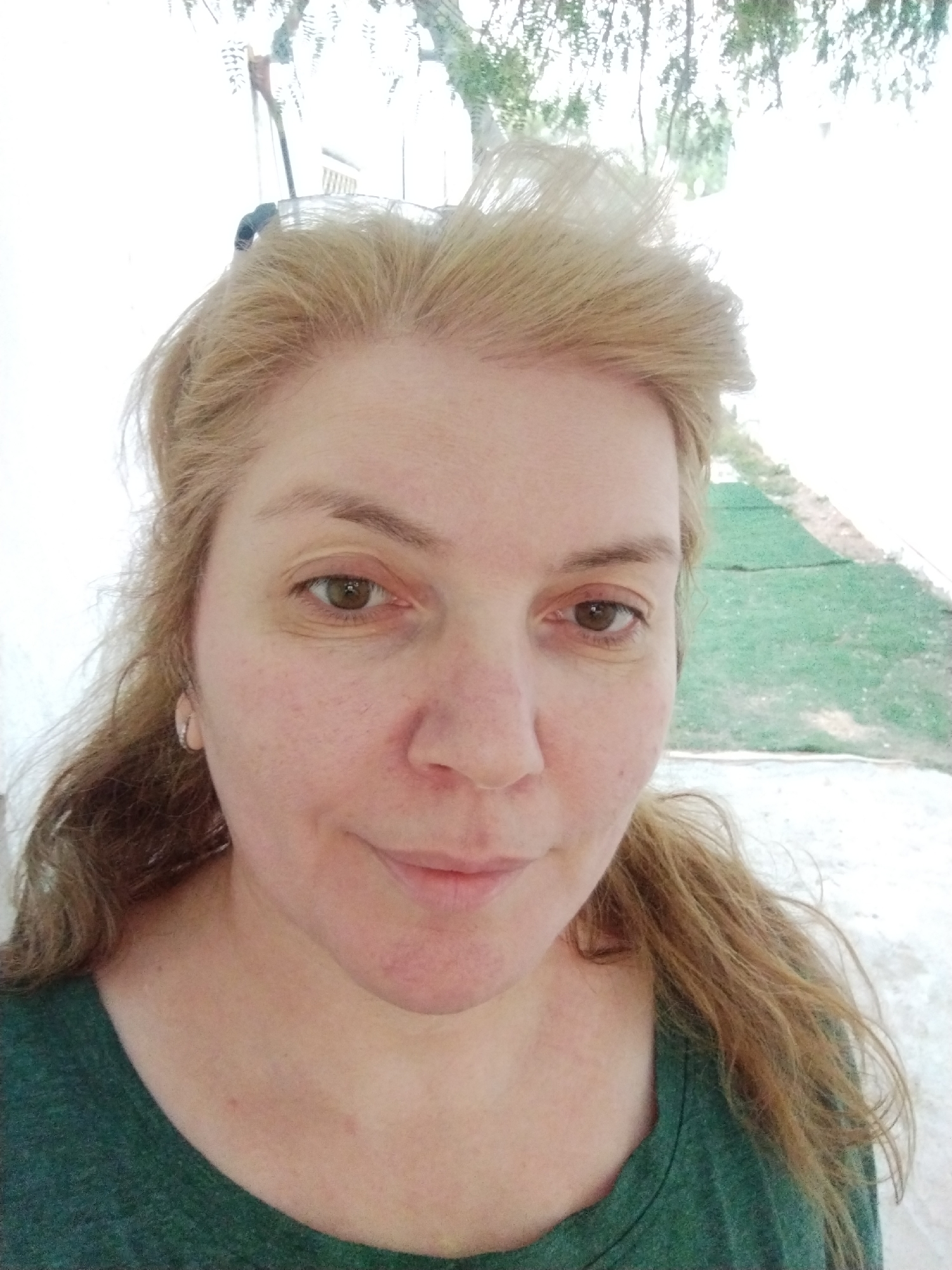A medium close-up shows a middle-aged woman with light skin and brown eyes. Her hair is blonde with darker roots and is styled off her face. She is wearing a green t-shirt. The background appears to be a white wall with some greenery and a window visible. The location in the city of Al Mas'oudi, United Arab Emirates is not possible to determine from this image.