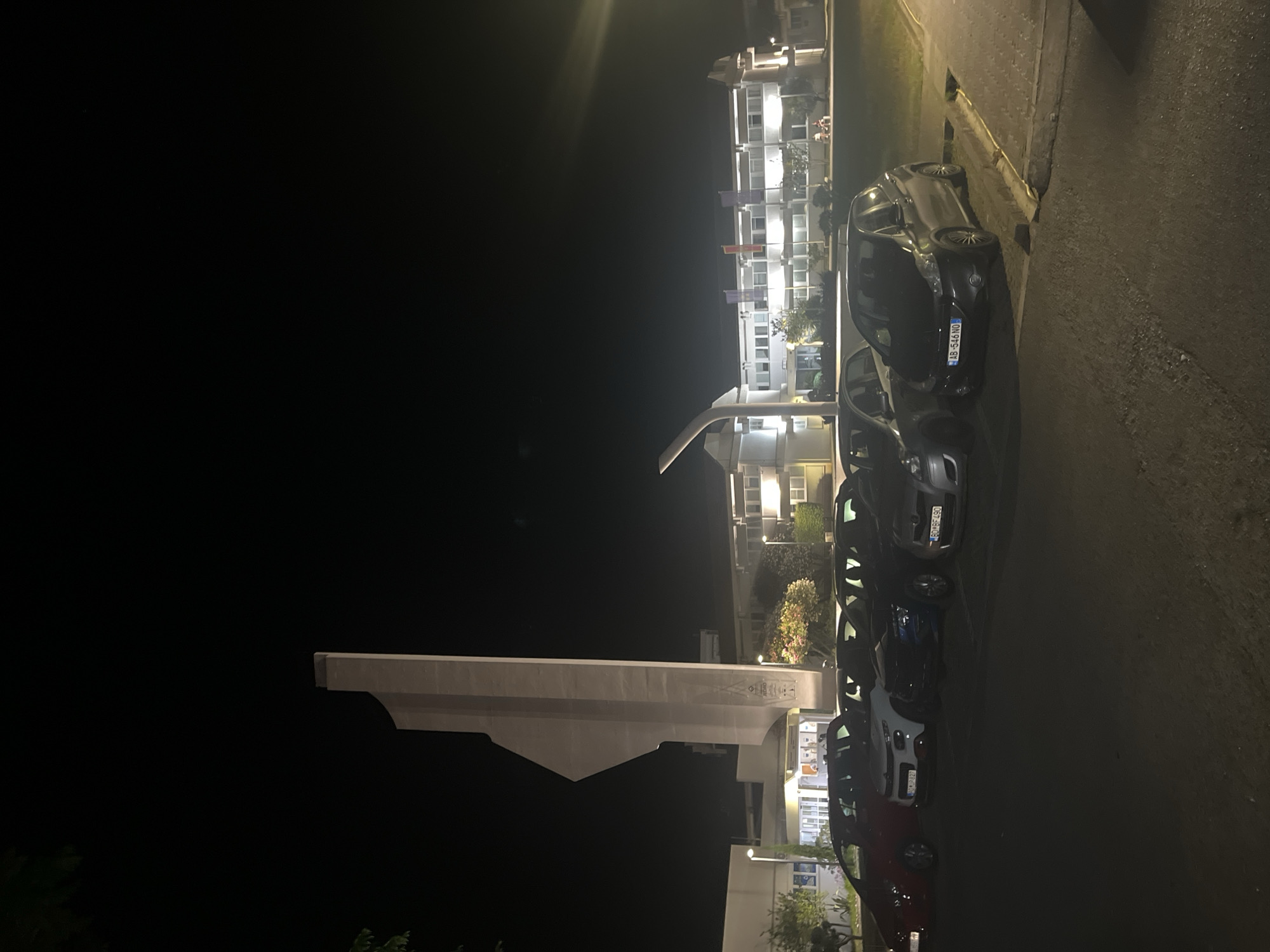 At night, a group of cars are parked in a lot in front of a modern, multi-story building with illuminated windows. The building appears to be a school or administrative complex, with flags displayed on its facade. To the left of the building, a tall, abstract concrete sculpture stands against the dark sky. The scene is captured from a low angle, with the foreground occupied by the parked vehicles, including a red car, a white Citroën, a blue hatchback, and a black Peugeot. The ground is asphalt, with yellow painted lines indicating parking spaces. The lighting suggests artificial illumination from the building and streetlights, casting a somewhat stark and dramatic atmosphere. No people are clearly visible in the immediate vicinity of the cars.